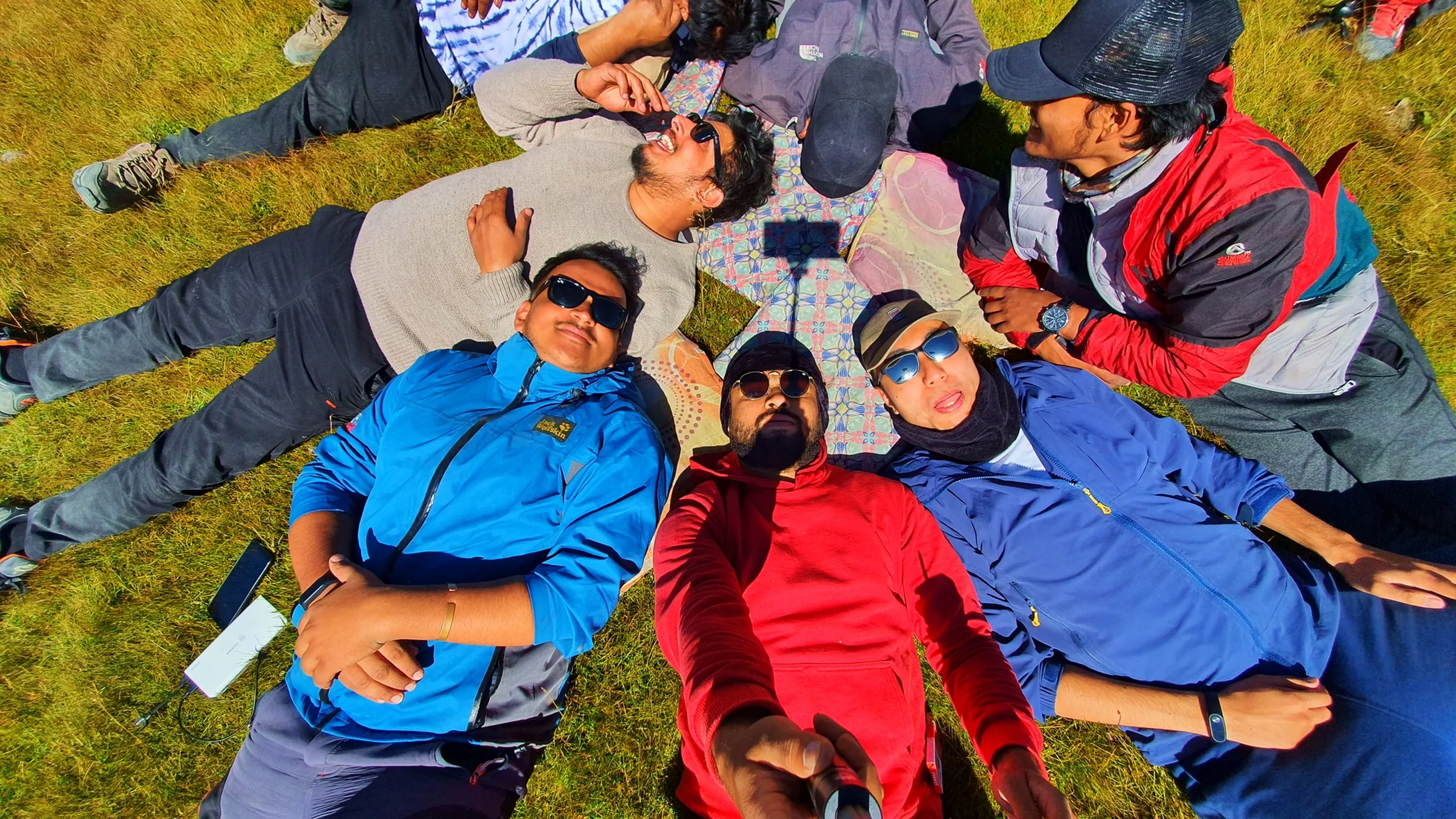 Overhead bird's-eye view of six trekkers lying flat on their backs in a circle on alpine grass in Manang Nepal, heads pointing inward like a human flower, all wearing colourful trekking jackets in blue, red, beige, and grey, grinning and laughing upward at the camera including one holding a selfie setup at the centre - Annapurna Circuit group rest, Nepal trekking fun, overhead selfie Himalayan meadow