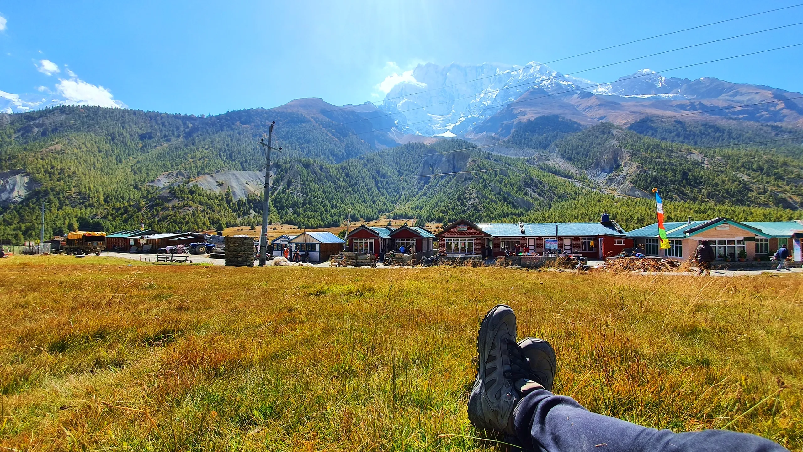 First-person POV rest shot lying on golden alpine grass in Manang Nepal, two black hiking boots crossed and pointed toward a row of small colourful guesthouse buildings with red, green and blue tin roofs, yaks and prayer flags visible in the distance, pine-covered hills rising behind and snow-capped Annapurna partially visible through bright sunlight - Manang Nepal guesthouse village, Annapurna Circuit rest stop, trekker POV Nepal