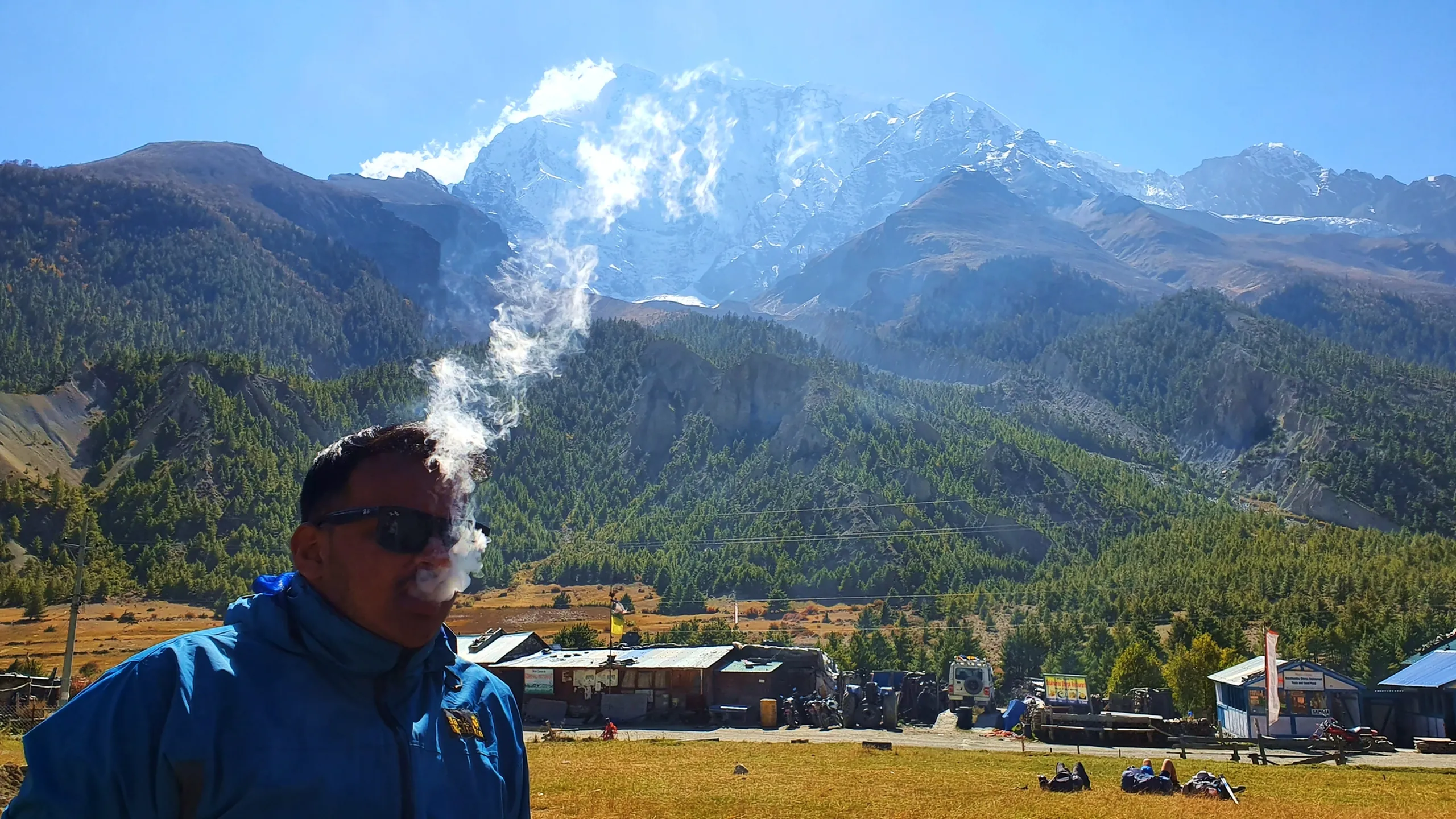 Male trekker in a blue jacket and sunglasses exhaling a visible plume of vapour or smoke against a dramatic backdrop of the snow-covered Annapurna massif rising above pine-forested Himalayan slopes, the small settlement of Manang visible in the valley below with vehicles and structures - Annapurna Circuit Manang village Nepal, dramatic mountain portrait, high altitude Nepal landscape