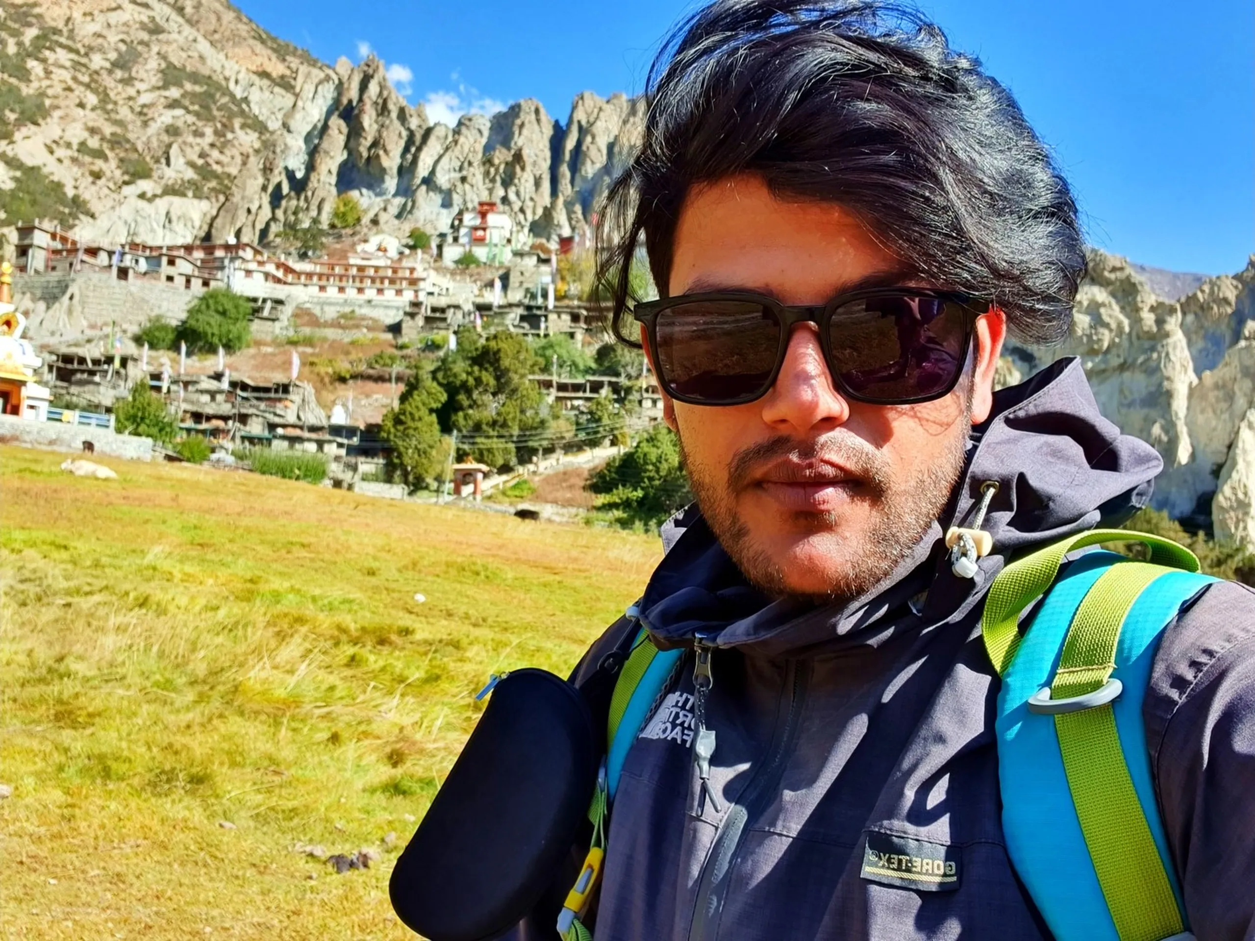 Close-up selfie of a male trekker in a dark North Face Gore-Tex jacket and dark square sunglasses with neon green backpack straps, wind-swept dark hair, looking seriously at the camera with Braga village - an ancient 800-year-old Himalayan settlement - stacked dramatically on eroded cliffs directly behind him, arid rocky spires above the village, clear blue sky overhead - Braga village Manang Nepal, ancient Himalayan village 800 years, Annapurna Circuit historical site