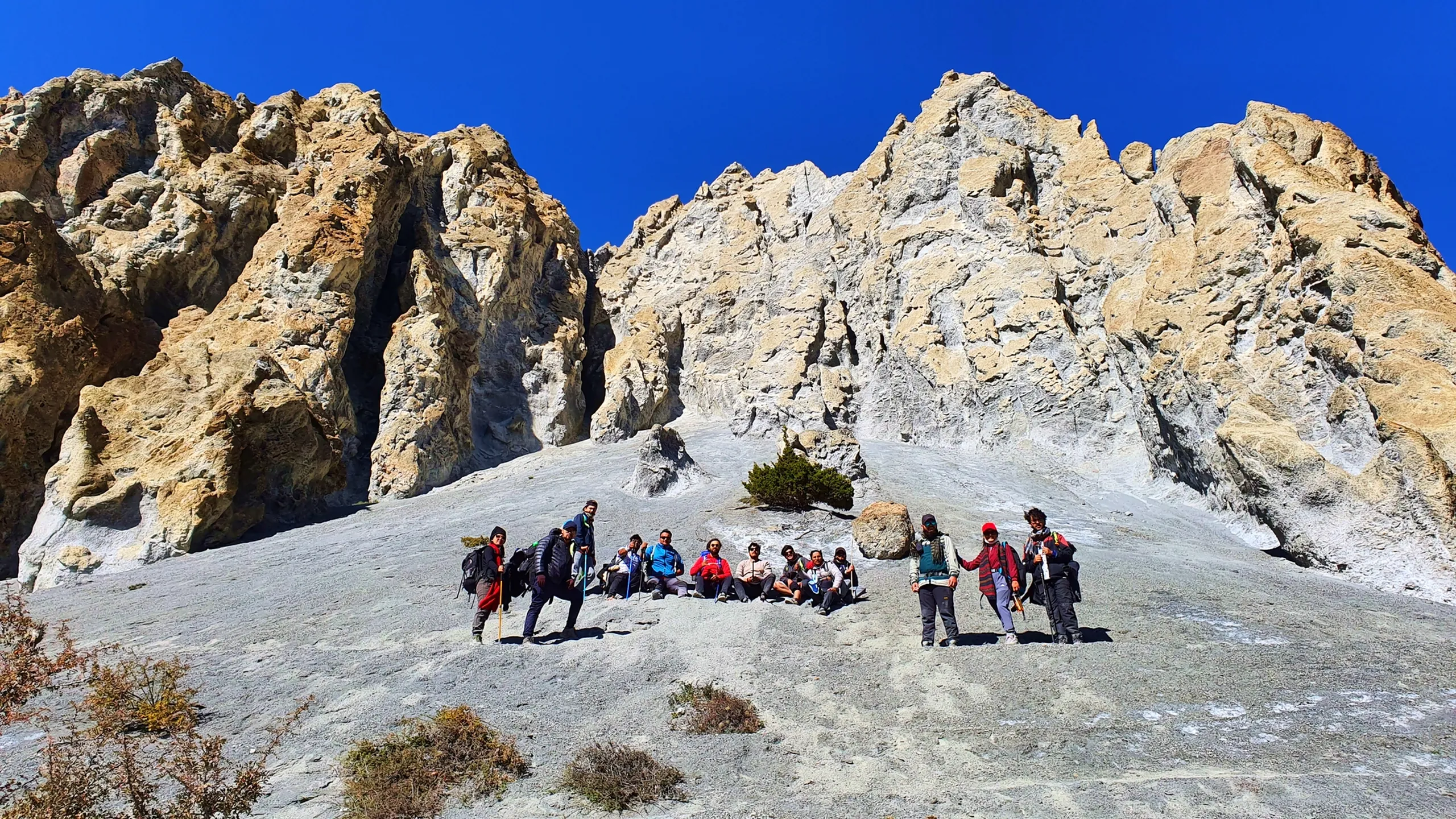 Wide-angle group photo of approximately twelve trekkers resting and posing on a steep smooth white limestone rock face flanked by dramatic jagged eroded rock towers, resembling the approach to Tilicho Lake Nepal; some sitting, some standing with trekking poles, all in colourful trekking gear, against a vivid deep blue sky - Tilicho Lake trail Nepal, Annapurna Circuit rocky terrain, high altitude rock climbing group Nepal