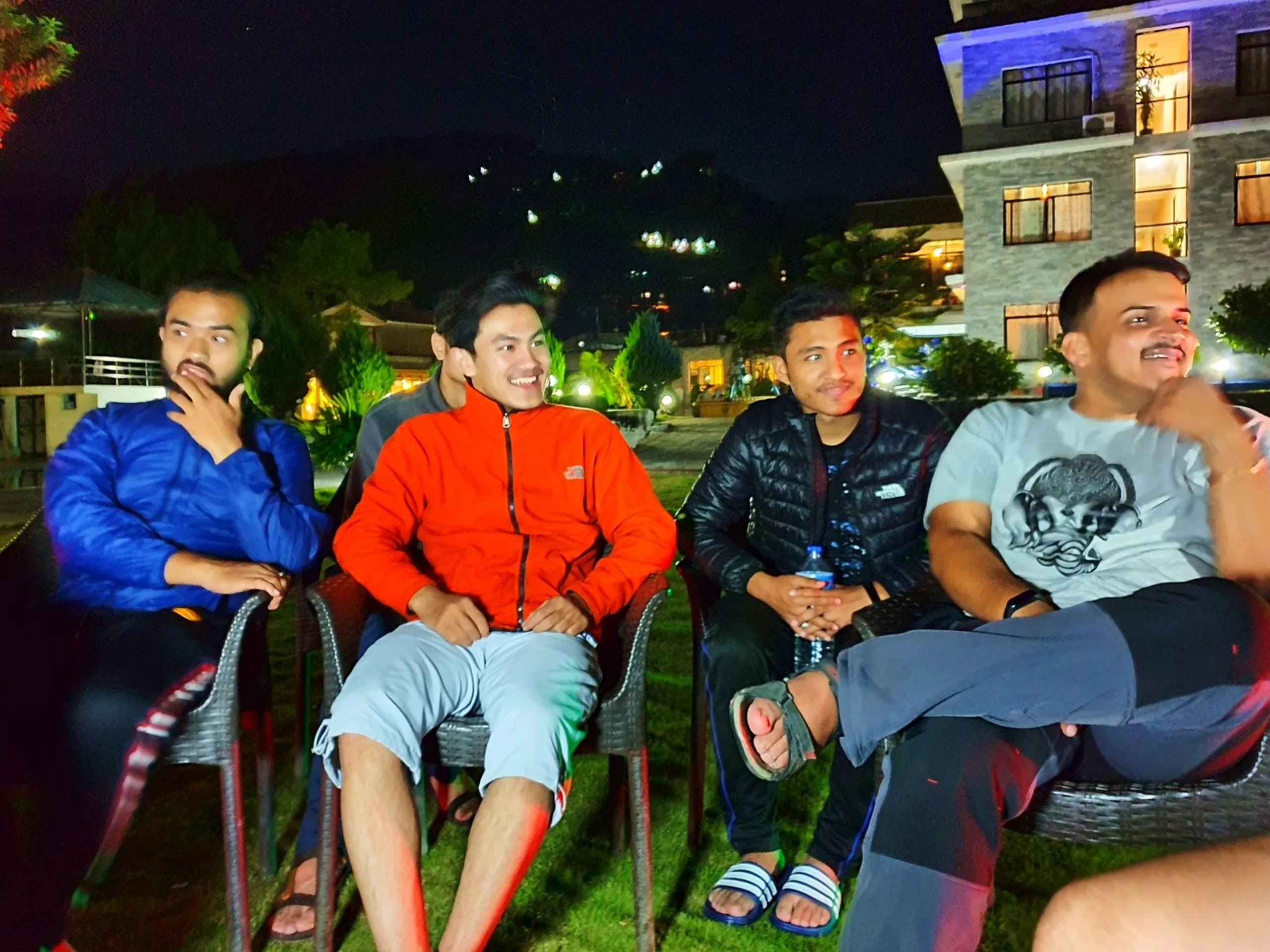 Four young male trekkers sit relaxed on outdoor garden chairs at night at a resort in Besisahar, Lamjung, Nepal - wearing puffer jackets and fleece layers in blue, red, and black - laughing and chatting under a dark sky lit by the resort's warm ambient garden lighting, with a multi-story stone-facade hotel building glowing warmly behind them and lush lit trees surrounding - a candid night scene capturing pre-trek excitement and friendship at a Nepal Himalaya base camp resort on the Annapurna Circuit