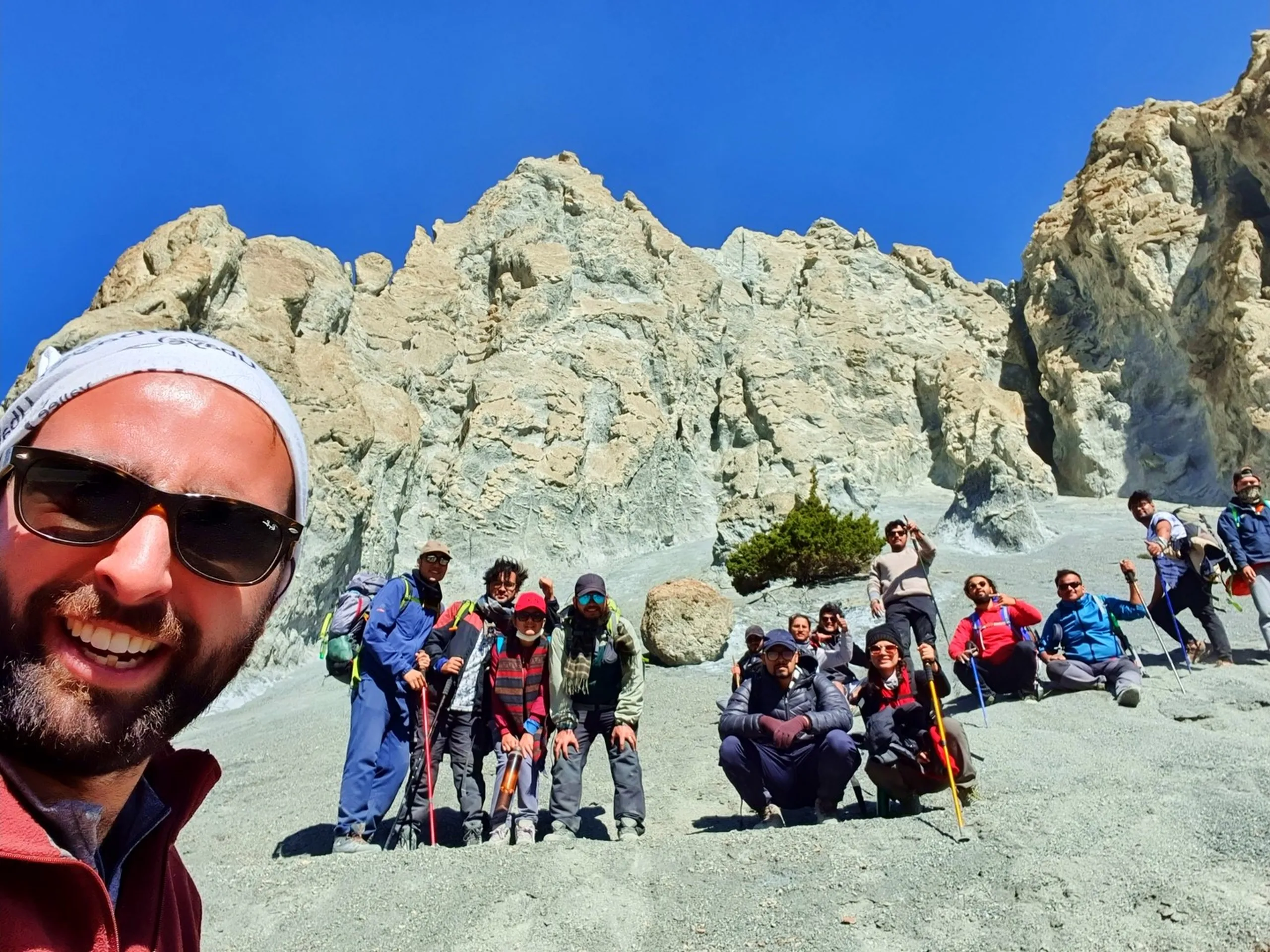Joyful group selfie taken by a smiling bearded foreign trekker in a white headband and sunglasses in the foreground, with approximately twelve Nepali and South Asian trekking companions arranged behind him on a white limestone rock face near Manang Nepal, trekking poles and colourful jackets visible, eroded rock pillars rising behind them into clear blue sky - international trekkers Nepal, Annapurna Circuit group selfie, meeting strangers trekking Nepal