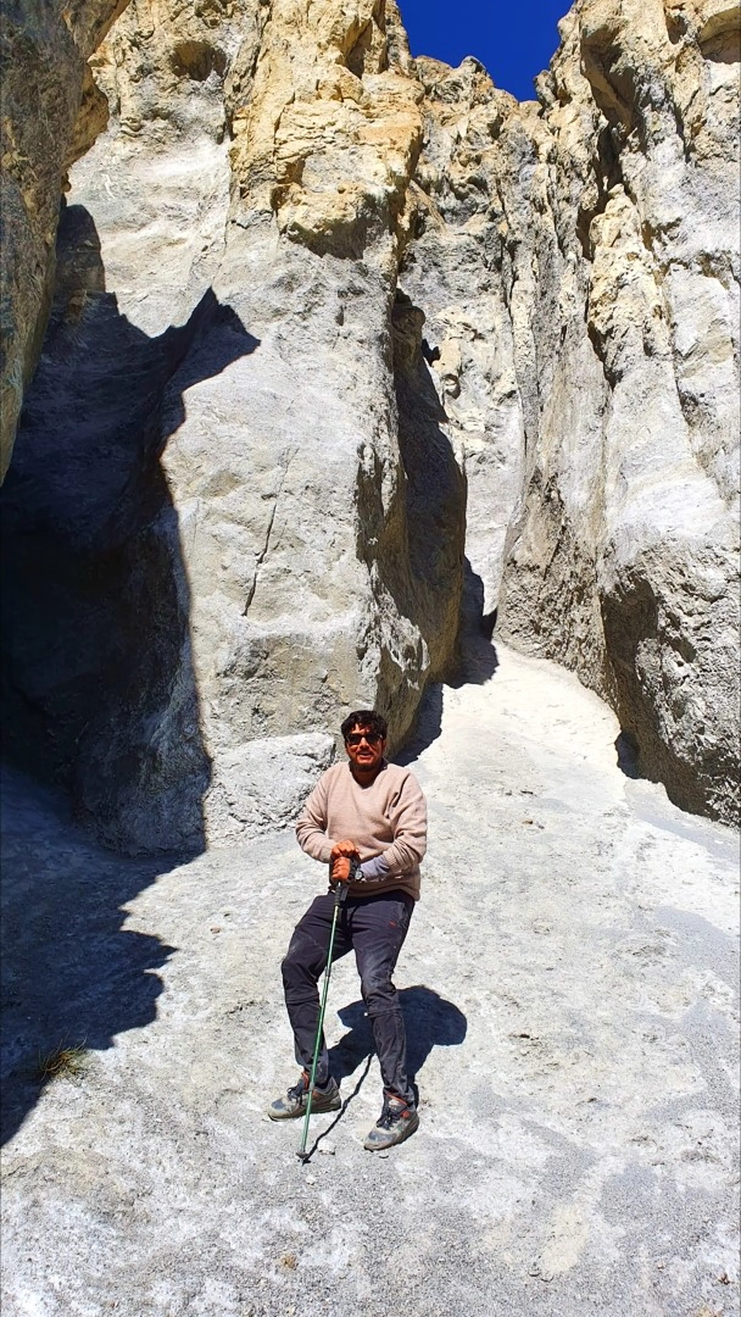Male trekker in a beige crewneck sweater and sunglasses standing confidently in a narrow gorge-like gap between two towering smooth white limestone rock walls near Manang Nepal, holding a trekking pole, the slot canyon opening to a bright blue sky above - Annapurna Circuit rock canyon Nepal, Manang geological formation, unique trekking terrain Nepal Himalaya