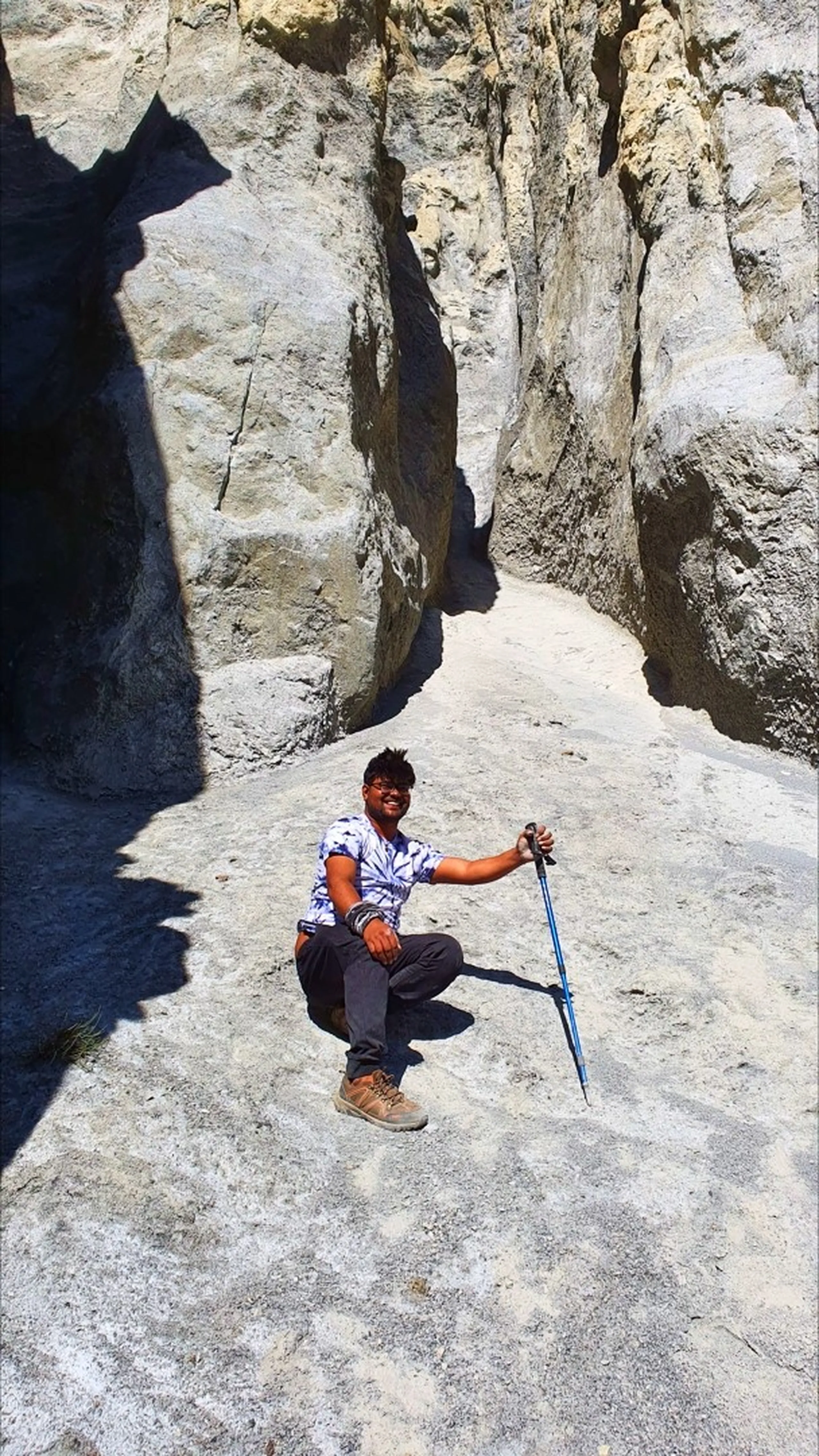 Smiling male trekker in a white and blue tie-dye t-shirt crouching on smooth white rock at the base of a dramatic narrow limestone slot canyon near Manang Nepal, holding a blue trekking pole, rock walls rising steeply on both sides, bright blue sky visible at the top of the slot - Annapurna Circuit rock formation Nepal, slot canyon Manang trekking, fun Nepal trek moment