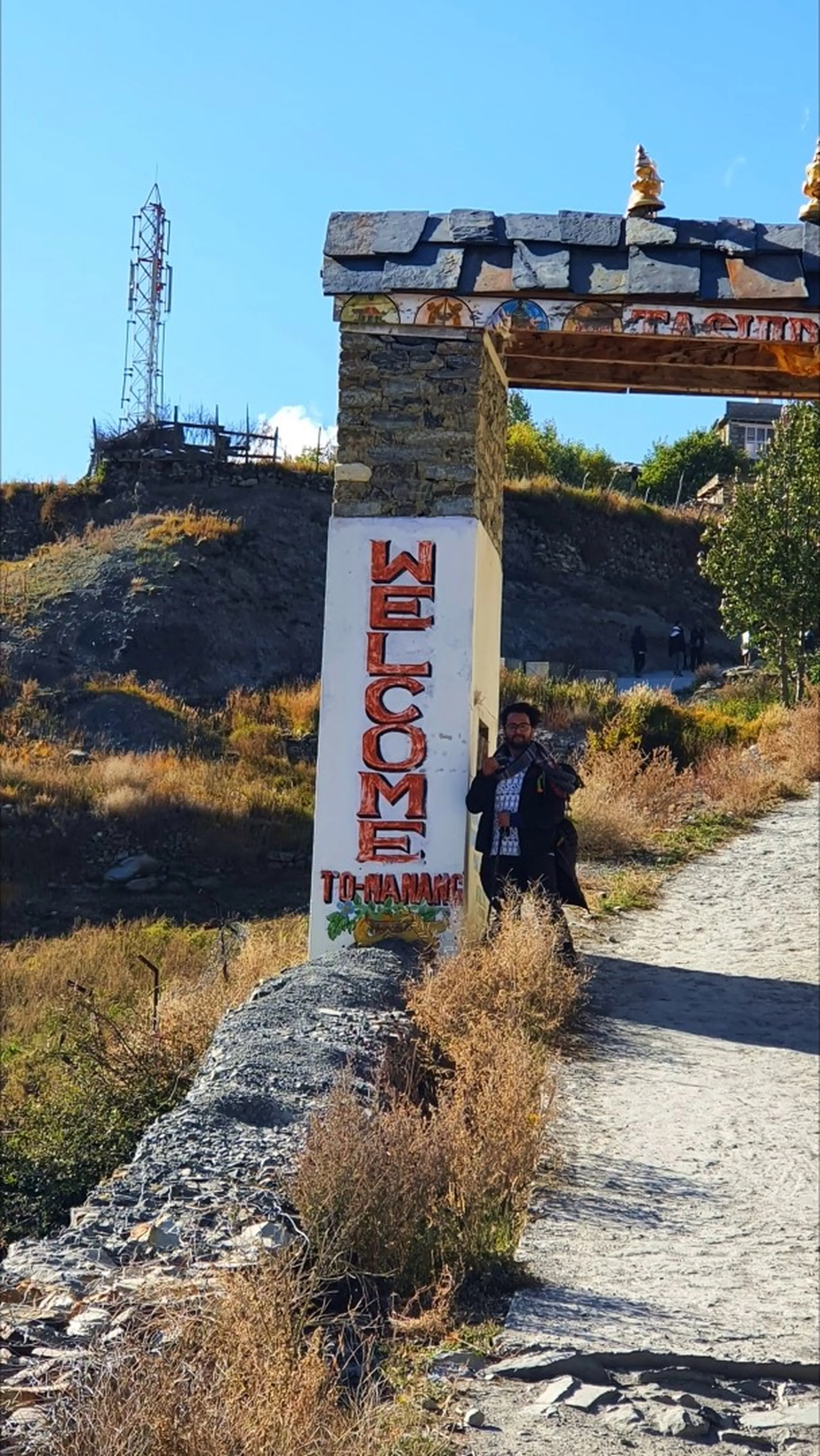 Male trekker leaning casually against the white pillar of a traditional stone-and-timber entry gate into Manang village Nepal, bold red hand-painted lettering reading "WELCOME TO MANANG" on the column, a golden stupa top visible above the gate arch, arid brown hillside and a telecommunications tower on the hill above, dusty gravel path leading through the gate - Welcome to Manang Nepal gate, Manang village entrance sign, Annapurna Circuit destination arrived