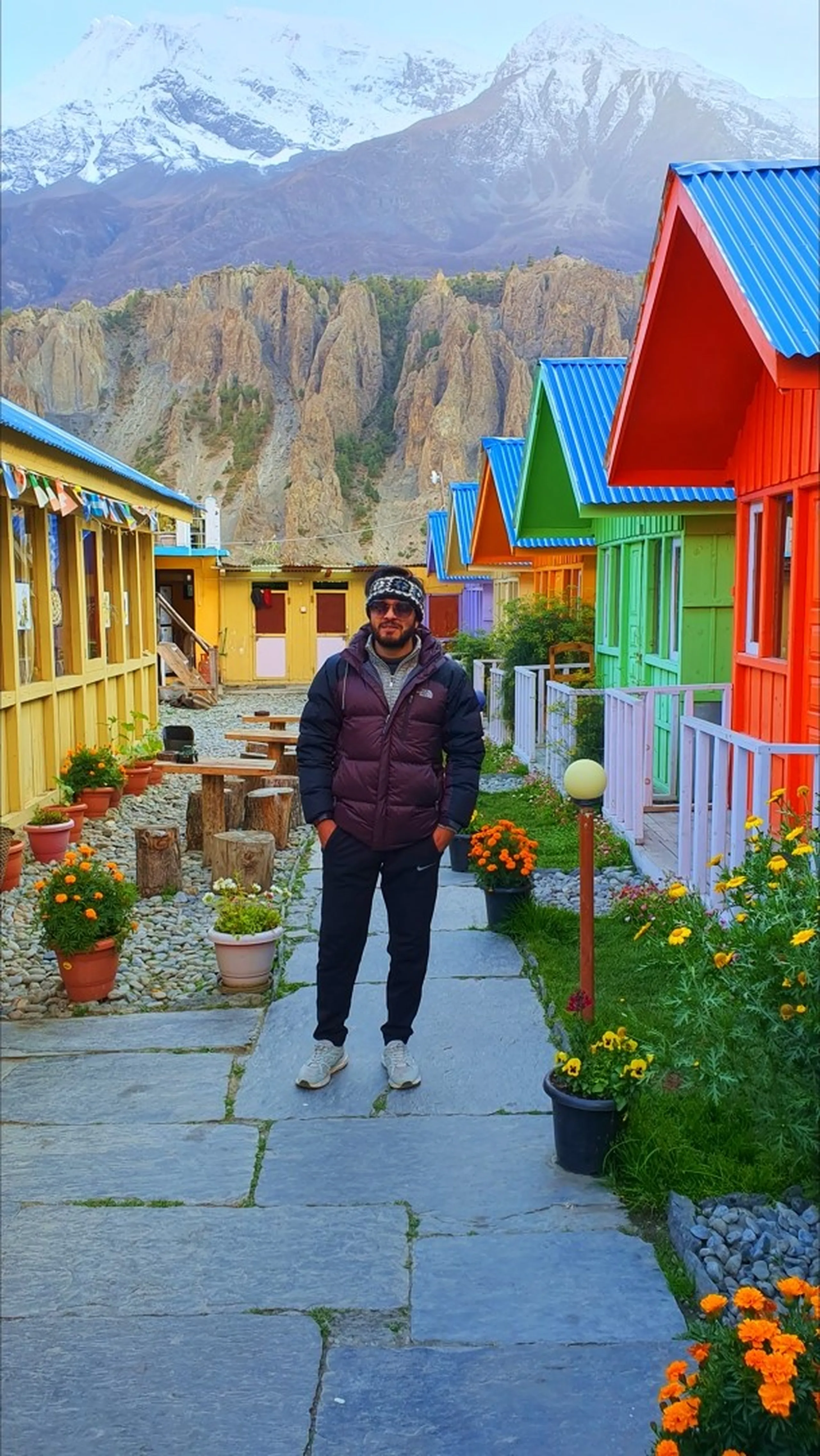 Male trekker in a dark North Face puffer jacket, printed headband and sunglasses standing in the middle of a stone-paved pathway flanked by a row of vibrant multicoloured wooden cottages - red, green, lime, blue, yellow and orange - at the famous Manang Alpine Home guesthouse in Manang Nepal, marigold flowers in pots lining the path, arid rocky cliffs and snow-dusted peaks visible behind - Manang Alpine Home Nepal, best guesthouse Manang Annapurna Circuit, colourful Nepal guesthouse