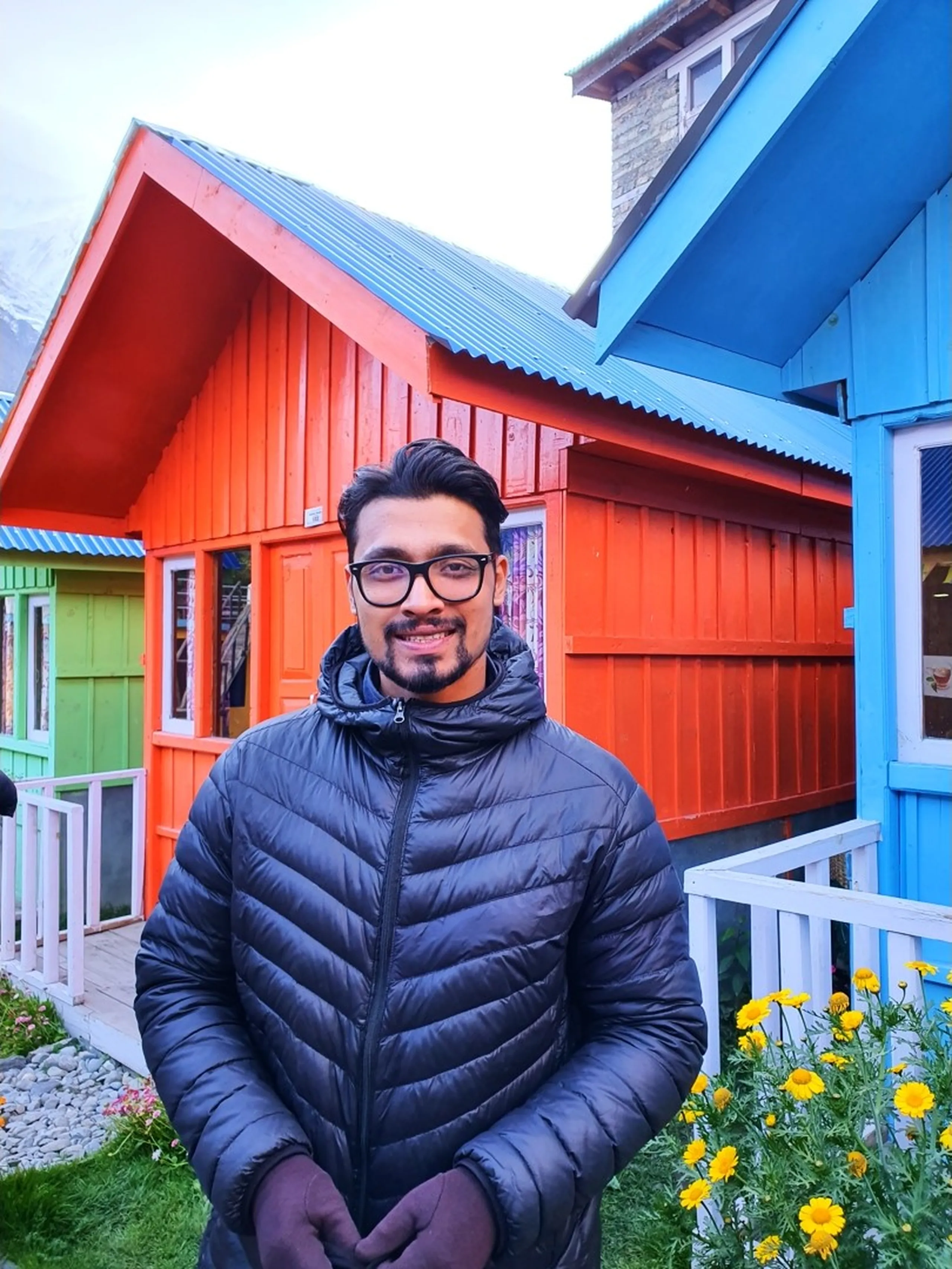 Smiling male trekker in a charcoal grey puffer jacket and black-framed glasses standing in front of a vivid orange-painted wooden cottage at Manang Alpine Home guesthouse Nepal, bright yellow marigold flowers in the foreground, a blue cabin visible to the right, overcast sky hinting at cooler mountain temperatures - Manang Alpine Home guesthouse, Annapurna Circuit accommodation Nepal, colourful mountain lodge Manang