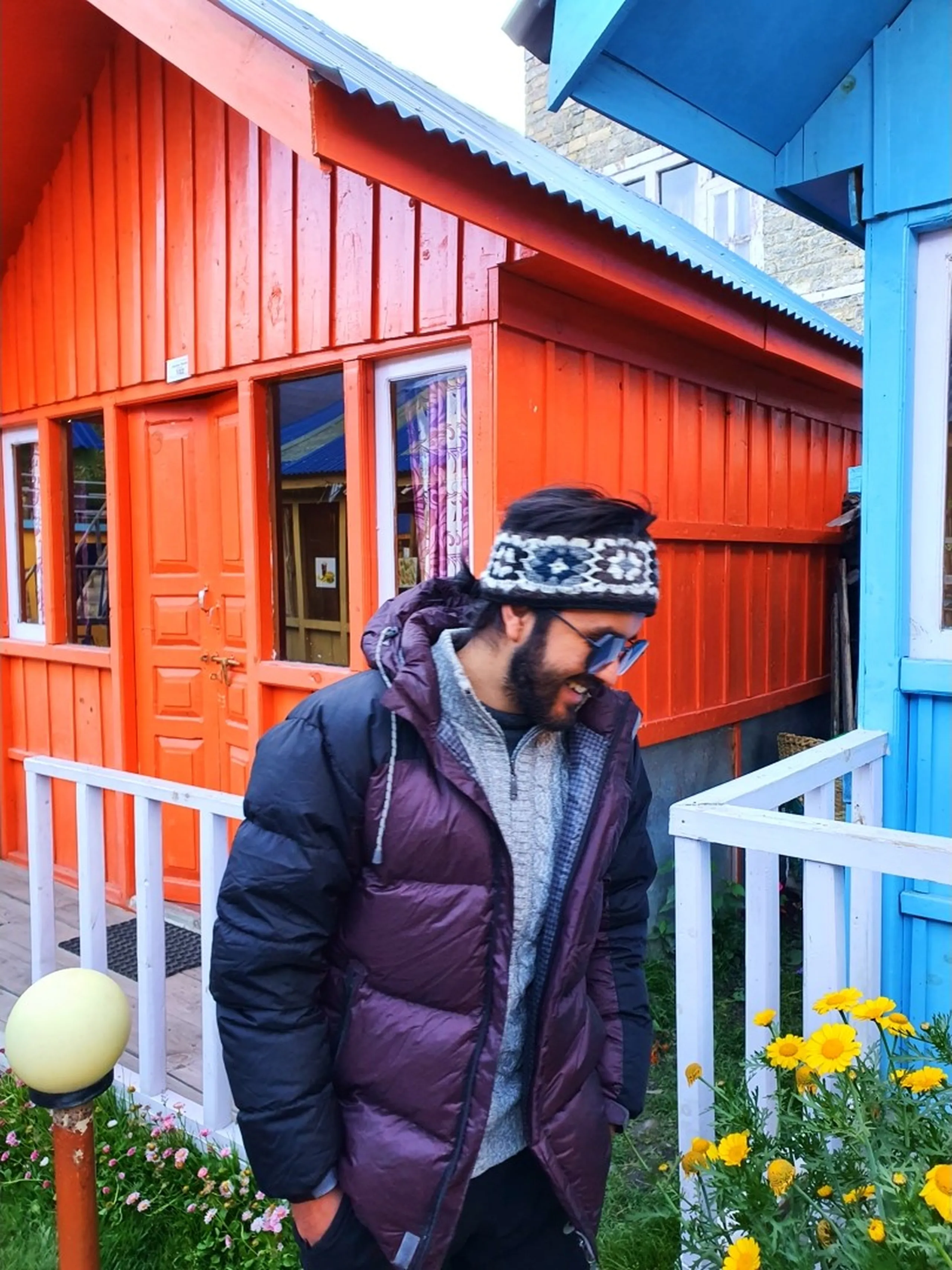 Candid moment of a male trekker in a dark purple puffer jacket and patterned headband looking down with a slight smile, standing outside the bright orange wooden cabin of Manang Alpine Home guesthouse in Nepal, yellow flowers at lower left, a round lamp post partially visible, blue cabin behind - Manang Alpine Home Nepal stay, Annapurna Circuit guesthouse candid, colourful cabin Manang village