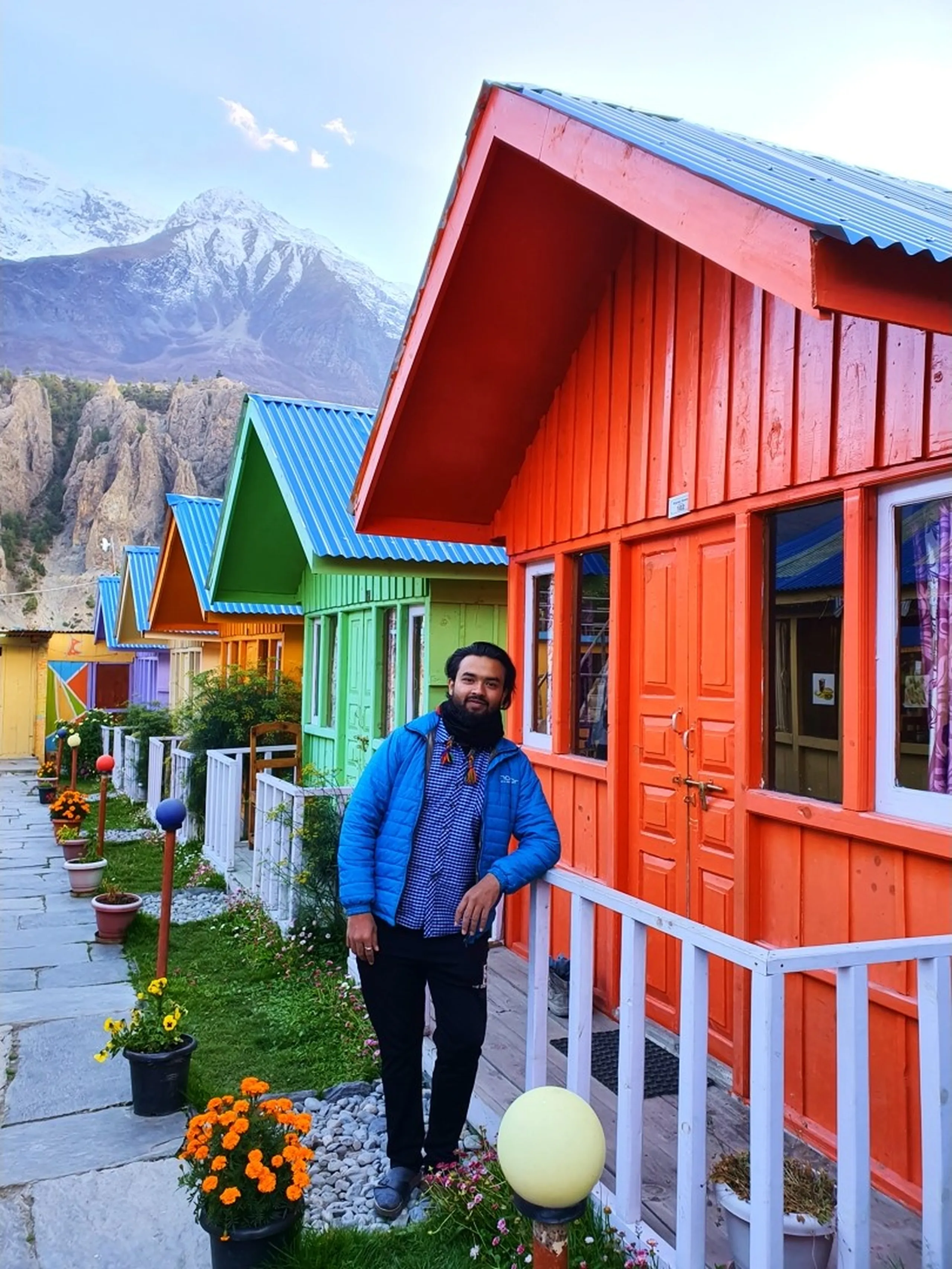 Male trekker in a blue puffer jacket over a blue checked shirt, standing beside the entrance of the bright orange cabin at Manang Alpine Home guesthouse Nepal, a full row of rainbow-coloured cottages receding into the background - red, green, lime, yellow, purple - with potted marigolds lining the stone path, eroded rocky cliffs and a snow-capped peak in the far background - Manang Alpine Home accommodation Nepal, Annapurna Circuit best stay, colourful guesthouse Manang