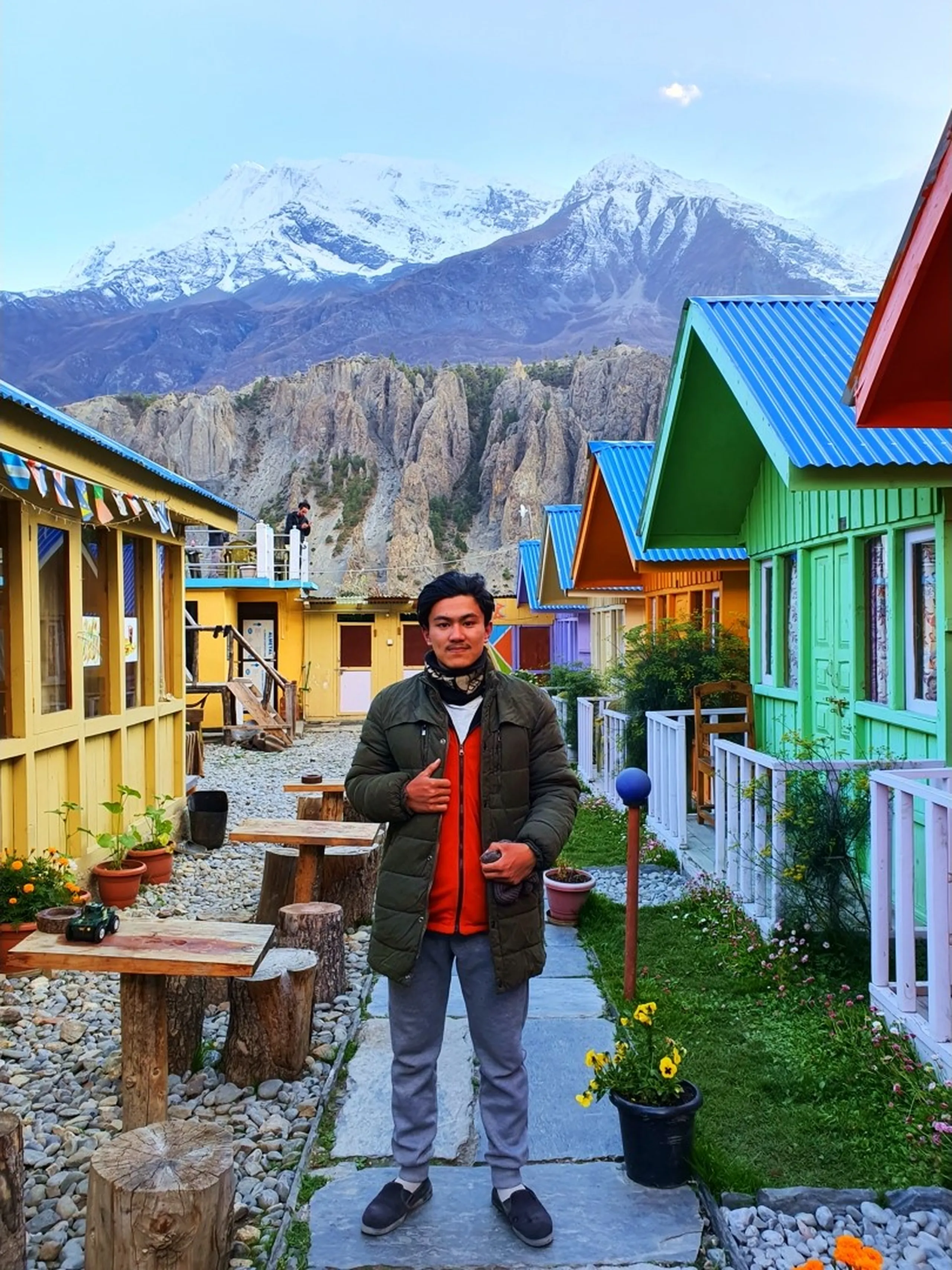 Young male trekker in an olive green puffer jacket over a red inner layer and grey joggers, standing in the central courtyard pathway of Manang Alpine Home guesthouse Nepal, bright green and red wooden cottage facades on either side, log-slab outdoor furniture visible, flower pots and a blue globe lamp on the path, snow-capped Annapurna mountains and eroded rock pinnacles filling the skyline in the background - Manang guesthouse courtyard Nepal, Annapurna Circuit colourful lodge, best hotel Manang Nepal