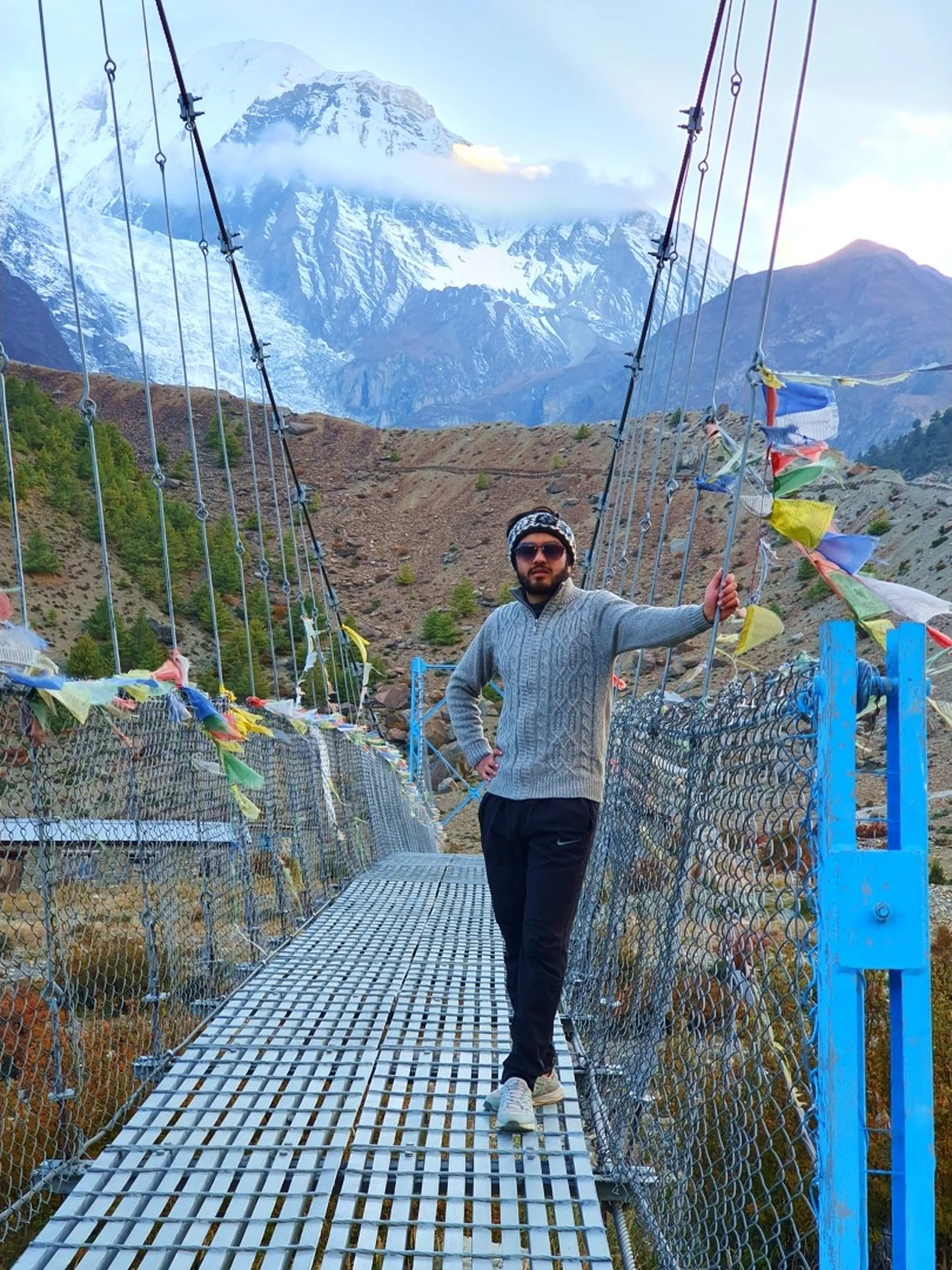 Male trekker in a grey cable-knit sweater, dark joggers, patterned woollen headband and pink-tinted sunglasses standing on a blue steel suspension bridge adorned with colourful Tibetan Buddhist prayer flags on both sides, one hand resting on the blue railing, the massive Gangapurna glacier and snowfield rising dramatically in misty clouds directly behind him - suspension bridge Manang Nepal, Gangapurna glacier lake trail, Annapurna Circuit bridge prayer flags