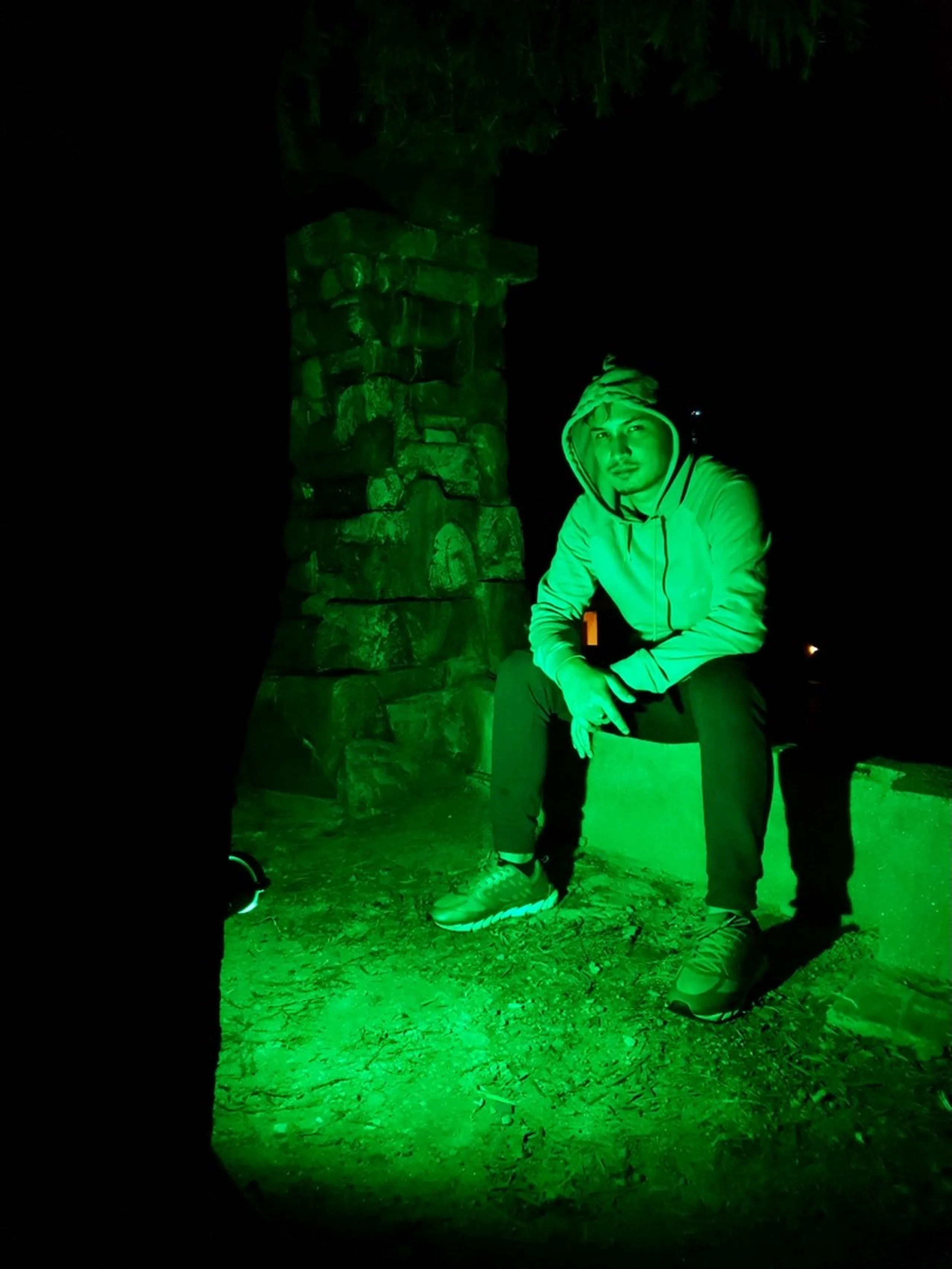 A lone young male trekker sits against a rustic stone pillar in near-total darkness, dramatically lit by an intense green LED light from below casting an eerie emerald glow across his hoodie, sneakers, and the earthen ground - his face partially illuminated, expression calm and contemplative - a strikingly moody and cinematic night photography composition at a Nepal trekking lodge, combining atmospheric lighting and raw stone architecture for a dramatic travel portrait