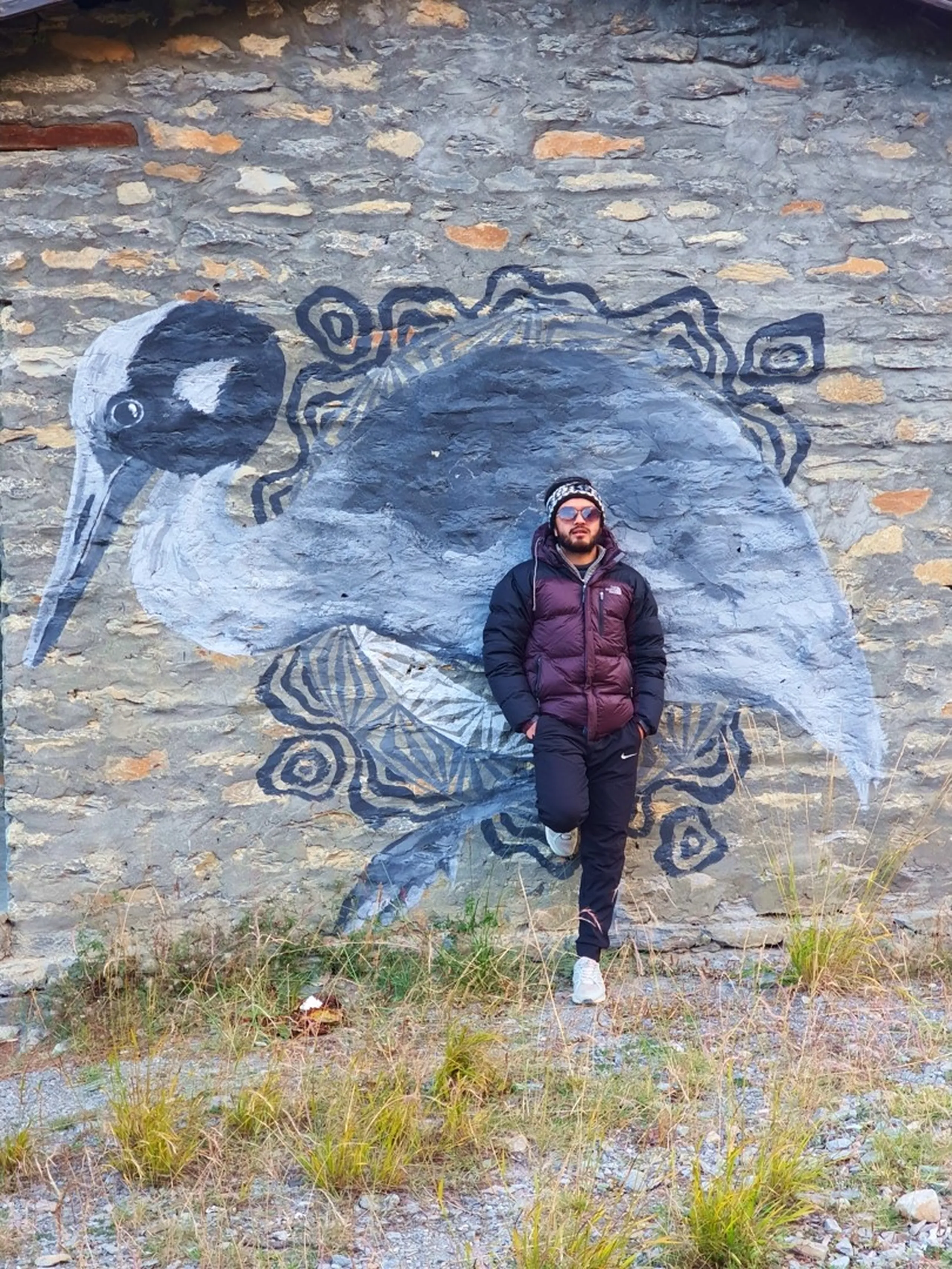 Male trekker in a dark maroon North Face puffer jacket, black Nike joggers, white trainers and patterned headband with blue mirrored sunglasses leaning against an old stone masonry wall painted with a large monochrome graffiti mural of a bird - resembling a Himalayan ibisbill or stork - with decorative circular patterns around it, dry grass in the foreground - Manang Nepal street art, Annapurna Circuit village mural, Nepal Himalayan bird graffiti wall art