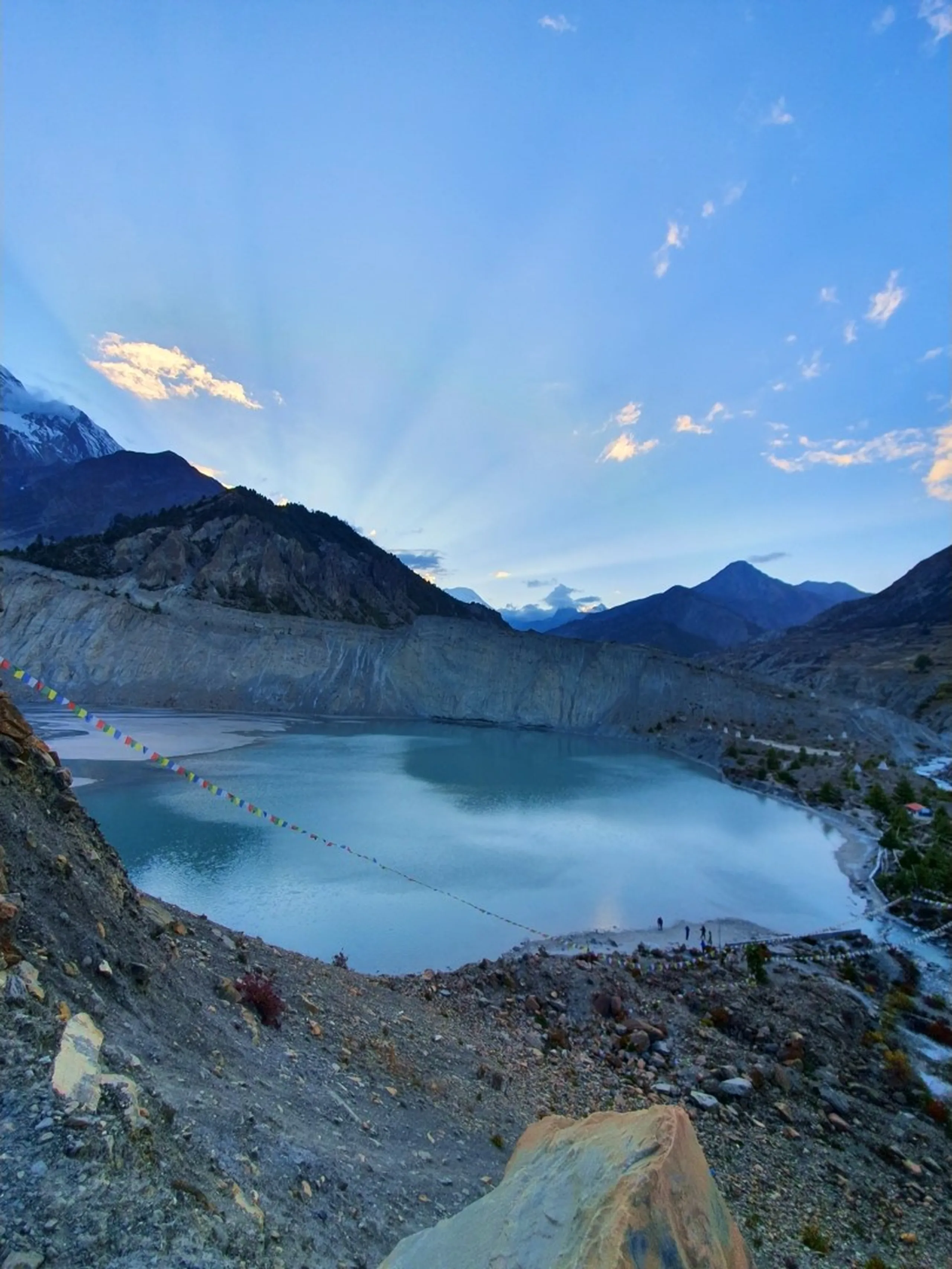 Breathtaking panoramic view of Gangapurna Glacier Lake in Manang Nepal at dusk, a wide turquoise glacial lake perfectly still and reflecting the pastel blue evening sky, a long string of colourful Tibetan Buddhist prayer flags strung diagonally across the rocky shoreline on the left, tiny human silhouettes standing at the far shore, surrounding Himalayan ridges in deep blue shadow, crepuscular rays bursting through clouds on the upper left - Gangapurna Lake Manang Nepal, Annapurna Circuit glacier lake, best lake view Nepal trekking