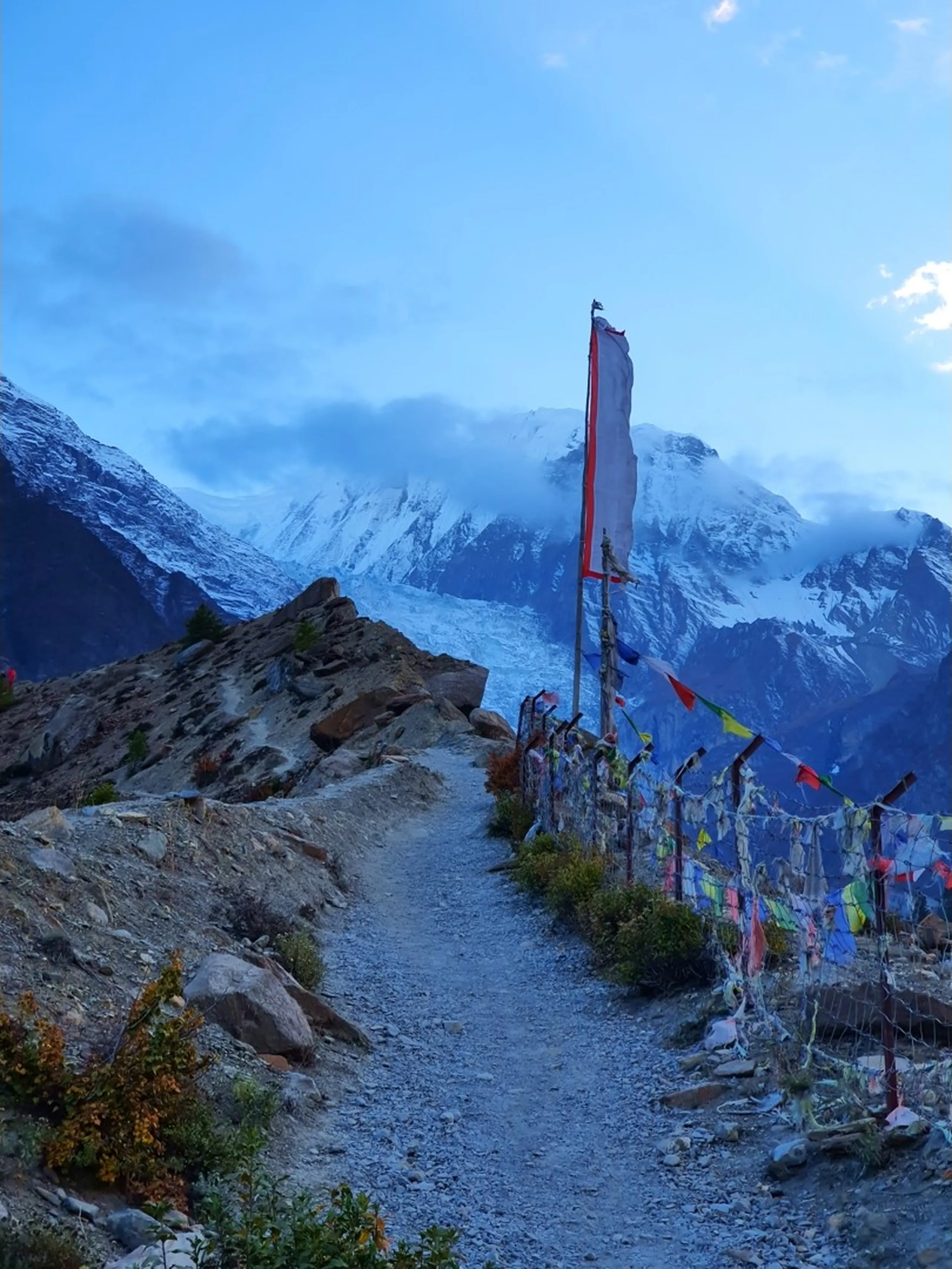 Atmospheric dusk viewpoint shot from the Gangapurna Glacier Lake trail in Manang Nepal, a narrow gravel path lined on the right by a wire fence adorned with densely tied multicolour Tibetan prayer flags including a large tall red and white flag on a pole, the massive Annapurna snowfield and glacier face emerging through evening clouds and blue light in the background - Gangapurna Lake viewpoint Nepal, Annapurna back side Manang, Himalayan prayer flags trail dusk