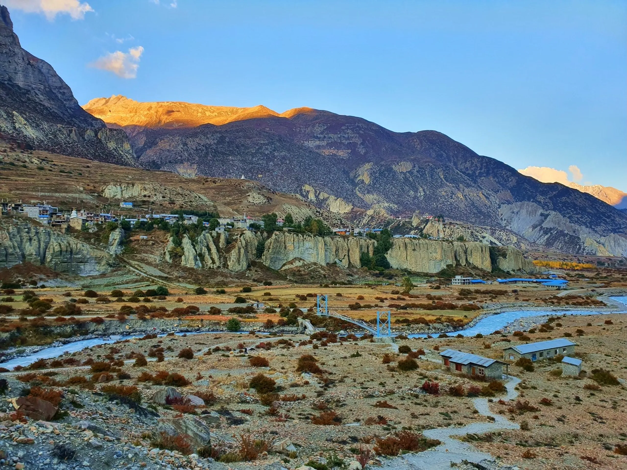 Golden hour landscape panorama of Manang village in the Annapurna Conservation Area Nepal, the cluster of whitewashed stone buildings perched above eroded rock spires and clay formations on a hillside, a blue suspension bridge crossing a glacial stream in the mid-ground, autumn red and orange shrubs across the valley floor, mountains catching last warm alpenglow above in a gradient from amber to violet sky - Manang village Nepal landscape, Annapurna Circuit village sunset, Himalayan village cliff Nepal