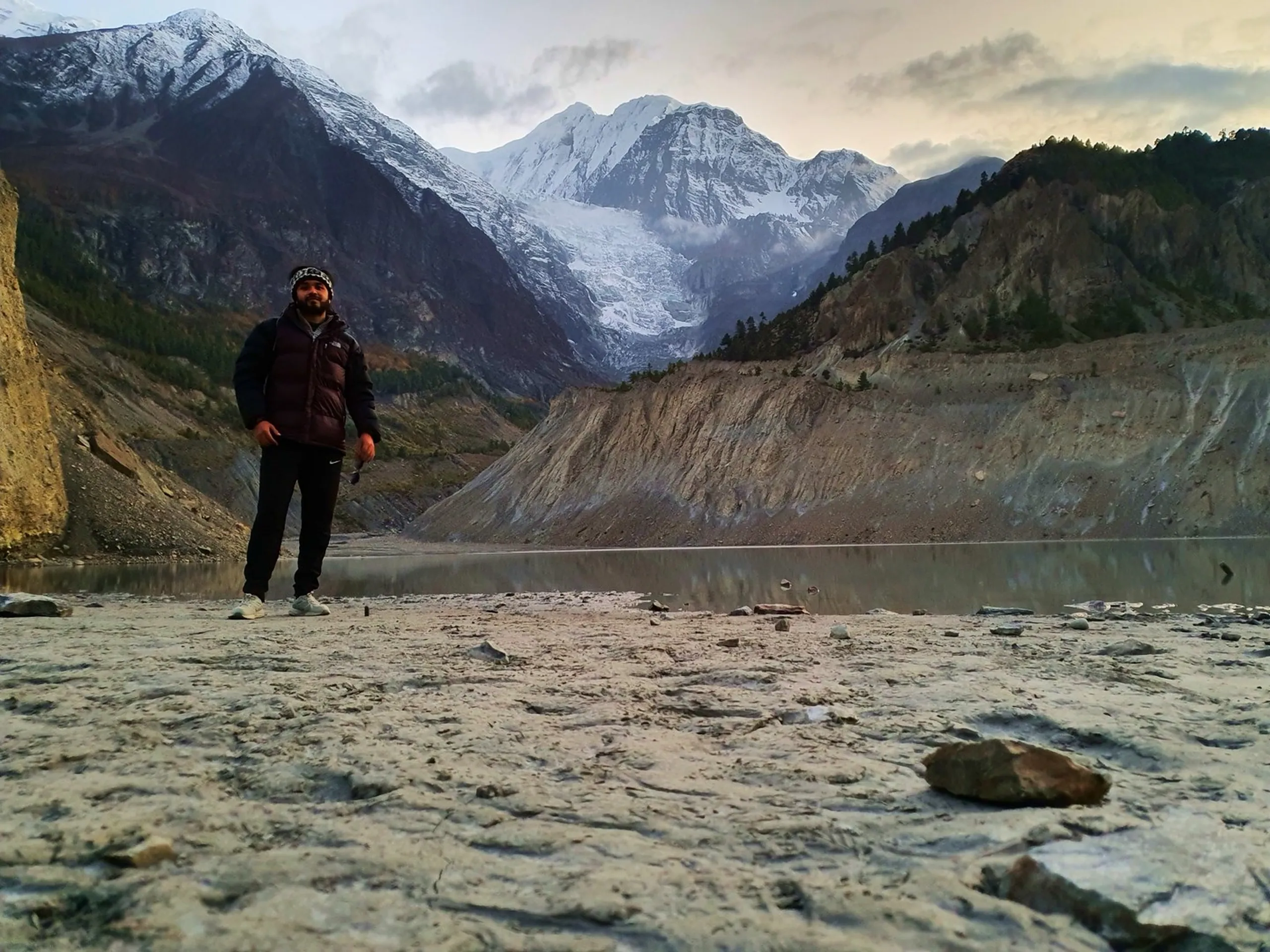 Male trekker in a dark maroon North Face puffer jacket and patterned headband standing alone on a flat glacial sand beach at the shore of Gangapurna Glacier Lake in Manang Nepal, moody overcast sky above, the Gangapurna glacier tongue flowing down between dark eroded ridges behind him, the still lake surface reflecting the pale sky - Gangapurna Glacier Lake Manang Nepal, Annapurna Circuit lake shore, glacier lake trek Nepal