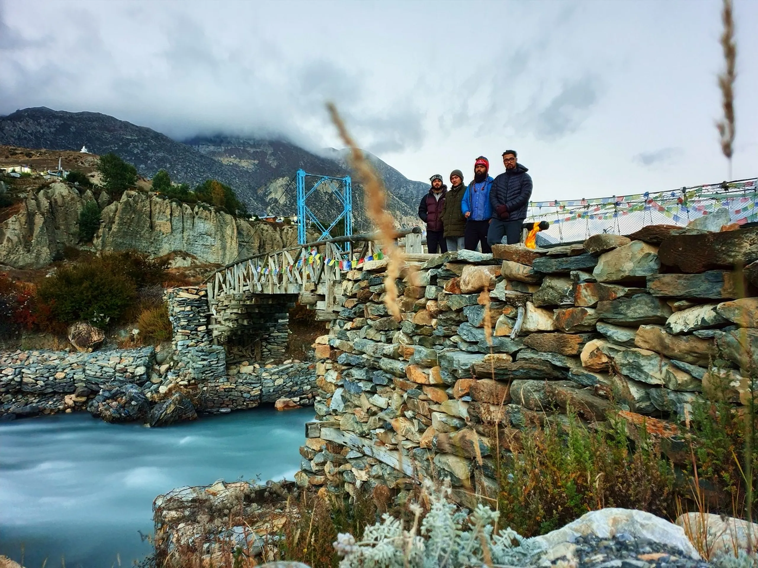 Four male trekkers posing together on a traditional dry-stone masonry bridge over a milky turquoise glacial river in Manang Nepal, a blue steel suspension bridge visible behind them, colourful prayer flags strung across both bridges, ancient Braga village stacked on eroded rock cliffs to the upper left, dark cloudy Himalayan sky above - Manang village bridge Nepal, Annapurna Circuit stone bridge, Marsyangdi river crossing Nepal trek