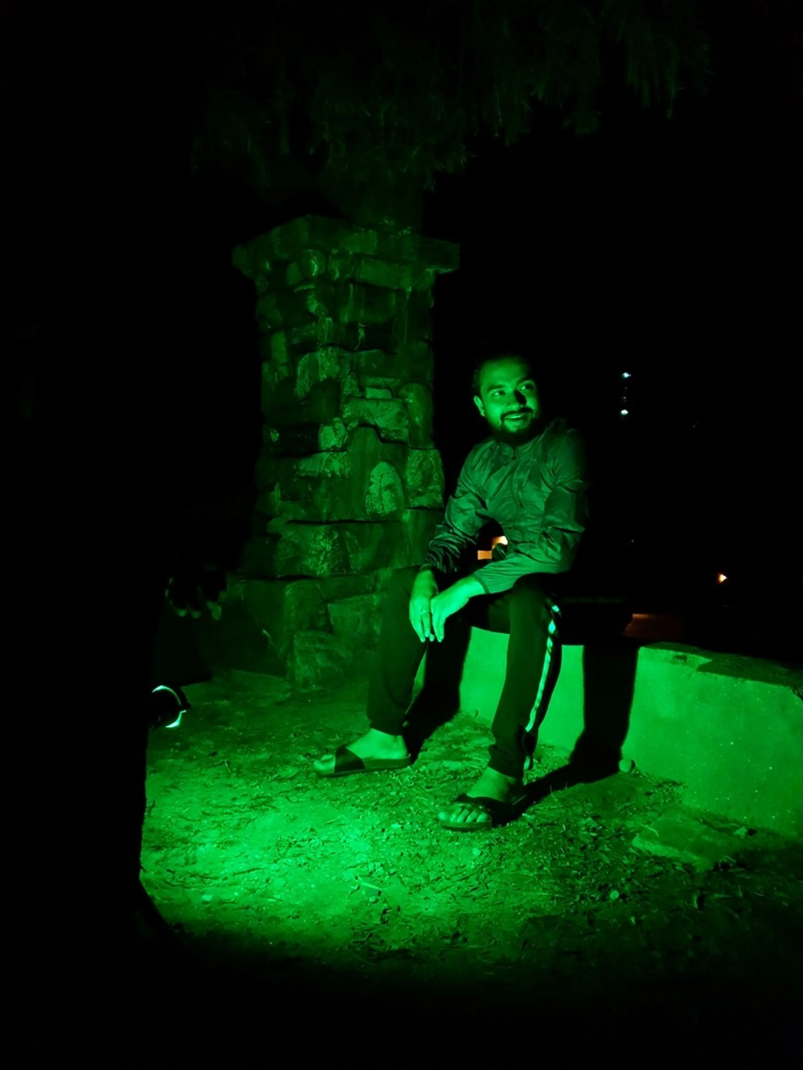 A young male trekker sits relaxed against a weathered stone pillar at night, bathed in vivid green LED light that illuminates his dark jacket, sandals, and the surrounding earth floor - smiling softly and looking slightly upward into the darkness - a water bottle tucked beside him - a moody and artistic night portrait at a Nepal mountain lodge, capturing the quiet reflective moments between trekking days on the Annapurna Circuit