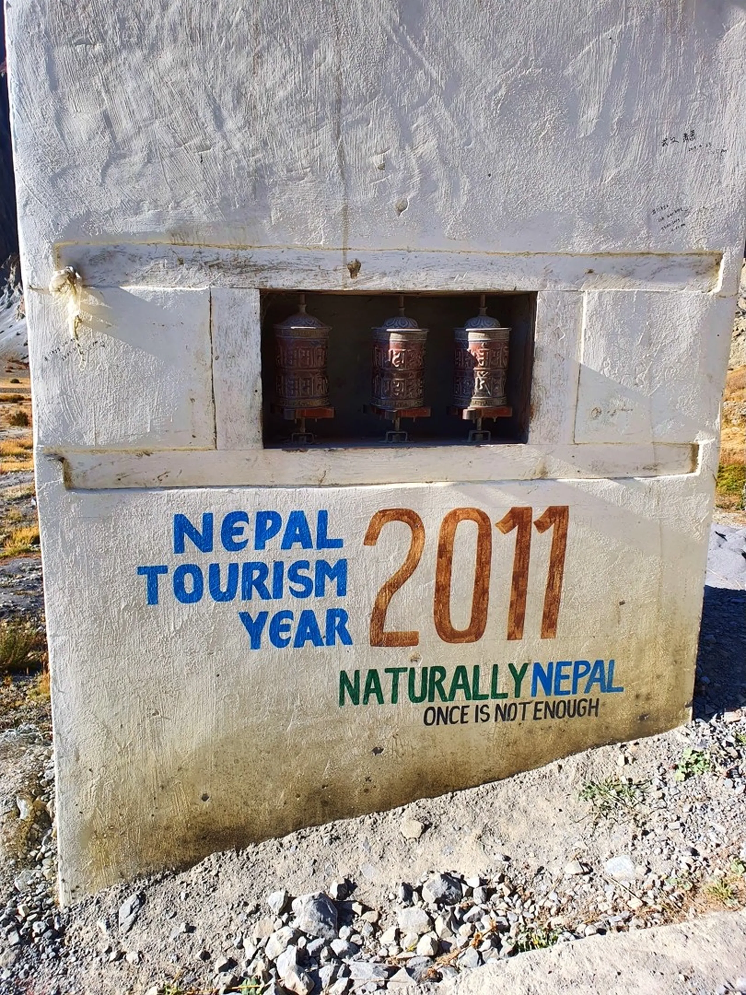 Male trekker in a blue zip jacket, cyan base layer, grey beanie and mirrored sunglasses standing confidently beside a weathered wooden "Ice Lake " trail signpost in Manang Nepal, other trekkers crouching with backpacks in the background adjusting gear, eroded rocky outcrops and a stone building visible behind, clear blue sky above - Ice Lake Manang Nepal trailhead sign, Annapurna Circuit Ice Lake trek start, Nepal high altitude lake trail