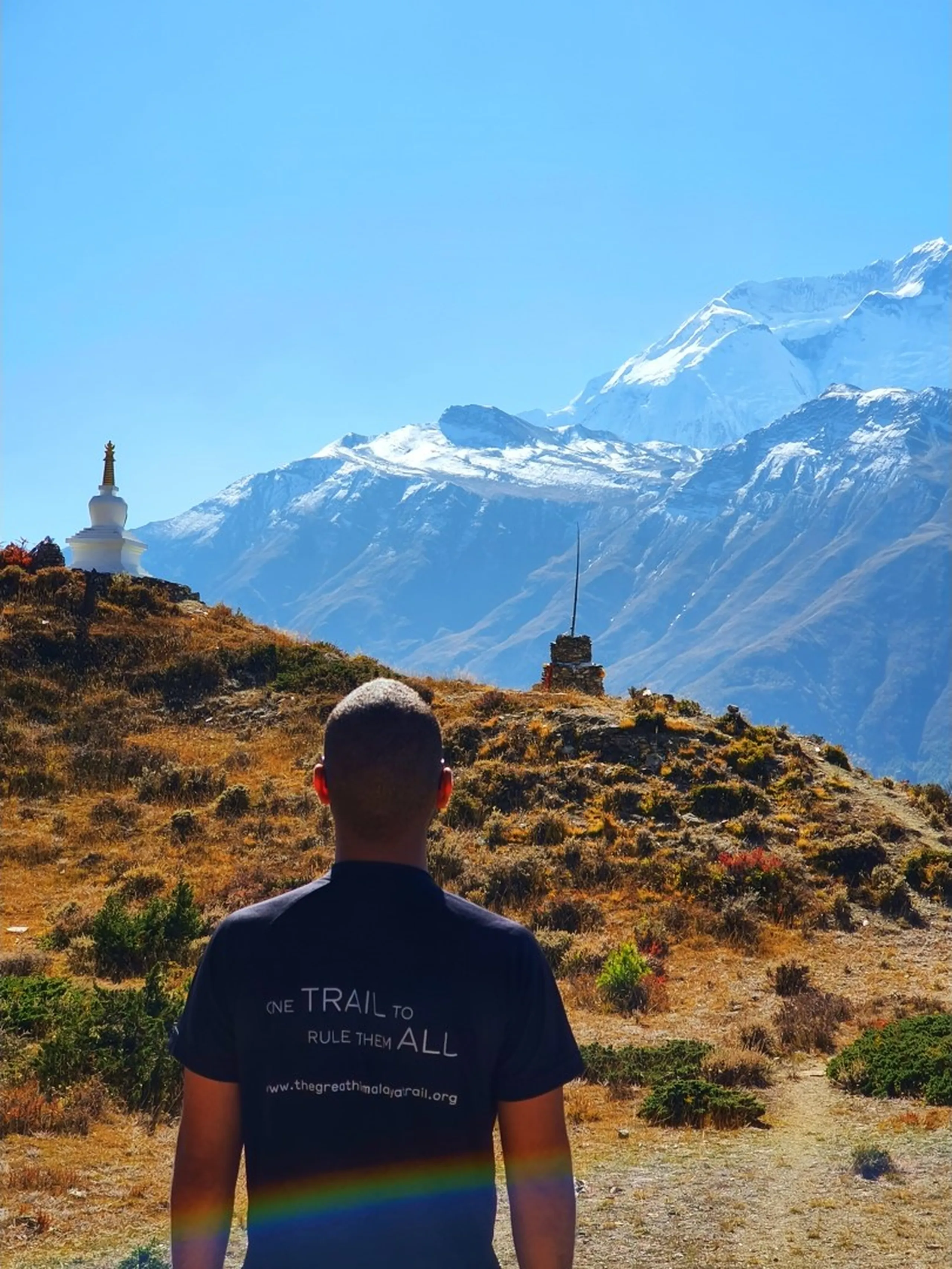 Male trekker in a blue checked puffer jacket standing on a high golden-grass ridge on the Ice Lake trail above Manang Nepal, beside a small cairn of balanced stones, a white Buddhist stupa visible on the adjacent hilltop, and the complete Annapurna and Gangapurna snowfields panorama stretching across the entire horizon with the Marsyangdi river valley far below and brilliant sunshine causing a lens flare - Ice Lake trek Manang Nepal viewpoint, Annapurna Gangapurna panorama, Himalayan high altitude ridge trail