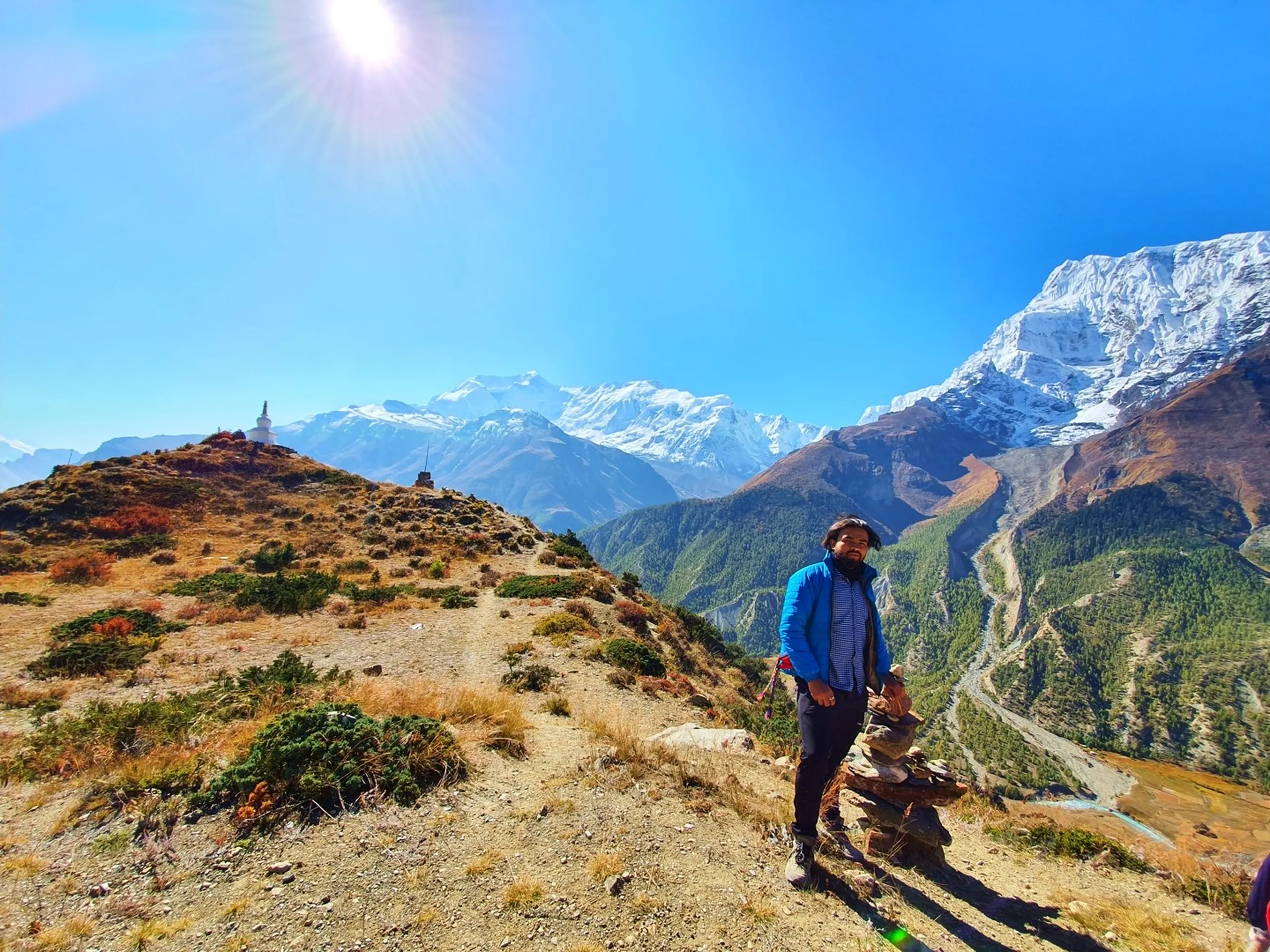 Male trekker in a blue Jack Wolfskin softshell jacket leaning on a trekking pole on a dry golden ridge on the Ice Lake approach above Manang Nepal, a stone shrine cairn with a flag pole behind him to the left, massive Annapurna II and Annapurna III snowfields filling the right half of the frame, a deep pine-forested valley below, under clear blue sky - Ice Lake trail Manang Nepal, Annapurna III viewpoint, high altitude trekking portrait Nepal