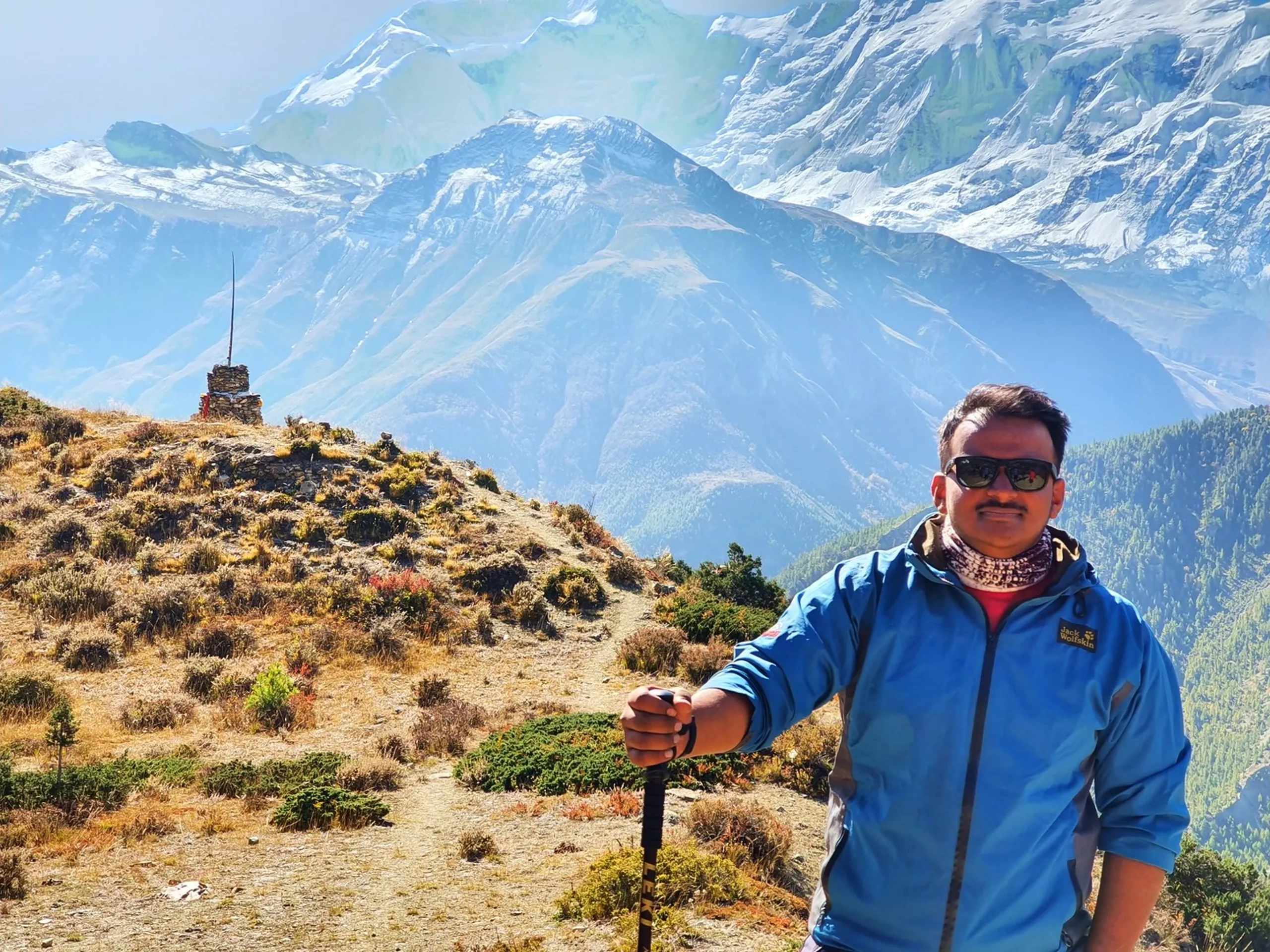 Male trekker in a dark navy puffer jacket and patterned woollen headband standing on the golden-grass Ice Lake ridge above Manang Nepal, looking directly at the camera with a calm expression, a small stone shrine cairn with a flag to his left, the enormous Annapurna snowfields and glacier faces filling the entire sky behind him - Ice Lake trail ridge Nepal, Annapurna Circuit high viewpoint, Manang high altitude portrait