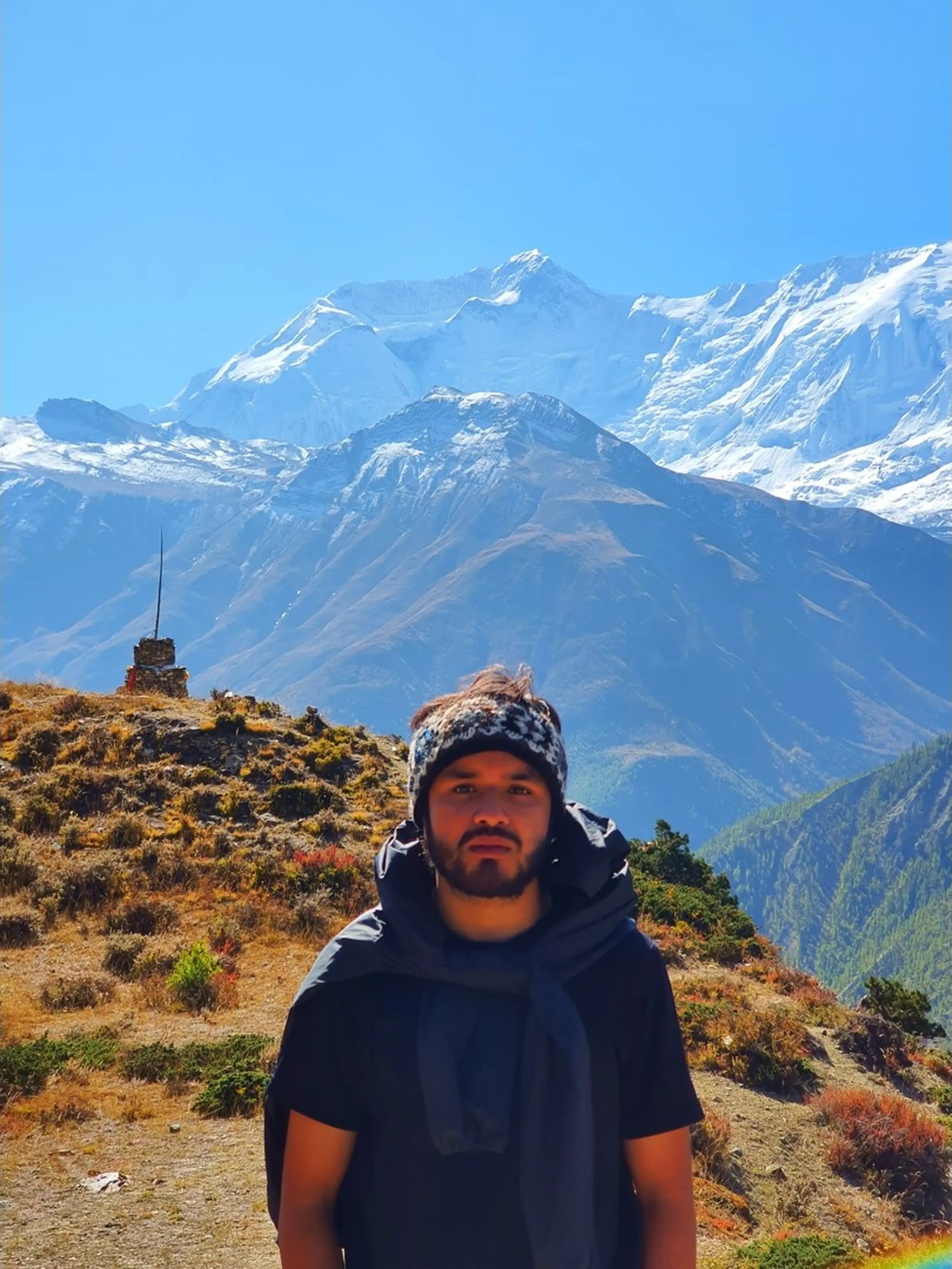 Male trekker in a dark jacket and patterned grey Chullo beanie standing on a high exposed ridge on the Ice Lake trail above Manang Nepal, wind moving through his hair, expression serious and contemplative, a stone shrine cairn just behind him to the left, and the spectacular Annapurna II massif completely dominating the background under a clear blue sky - Ice Lake trek Nepal portrait, Annapurna high altitude ridge, Manang trek solo male trekker
