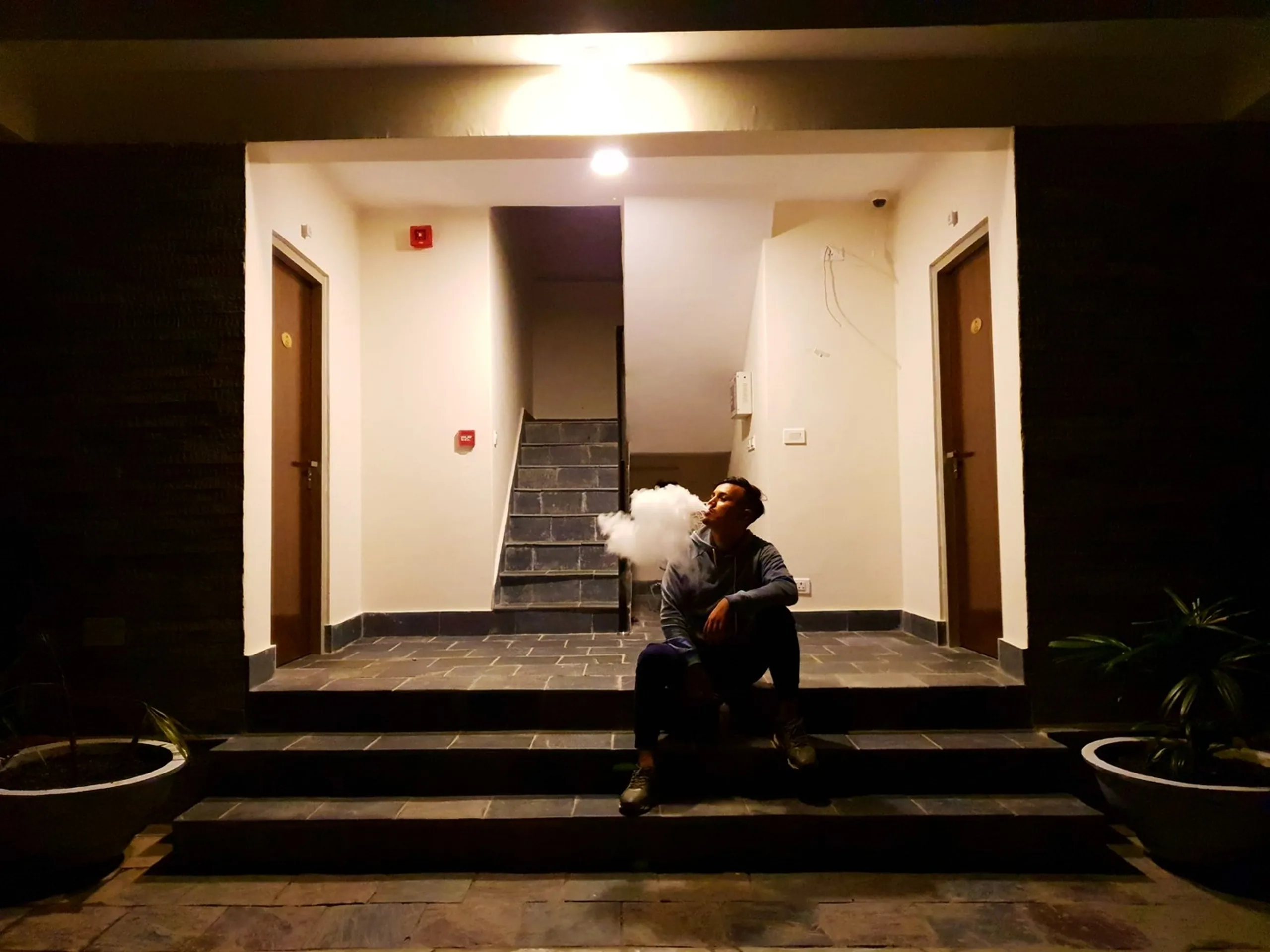 A young male trekker sits alone on stone steps outside a well-lit hotel building at night, exhaling a massive white vape cloud that billows dramatically in the warm overhead light - centered symmetrically between two wooden doors on either side of a staircase, with potted plants framing the entrance - a cinematic and moody night travel portrait at a Nepal trekking resort in Besisahar or Manang region, capturing the solitary late-night atmosphere of long trekking expeditions in the Himalayas