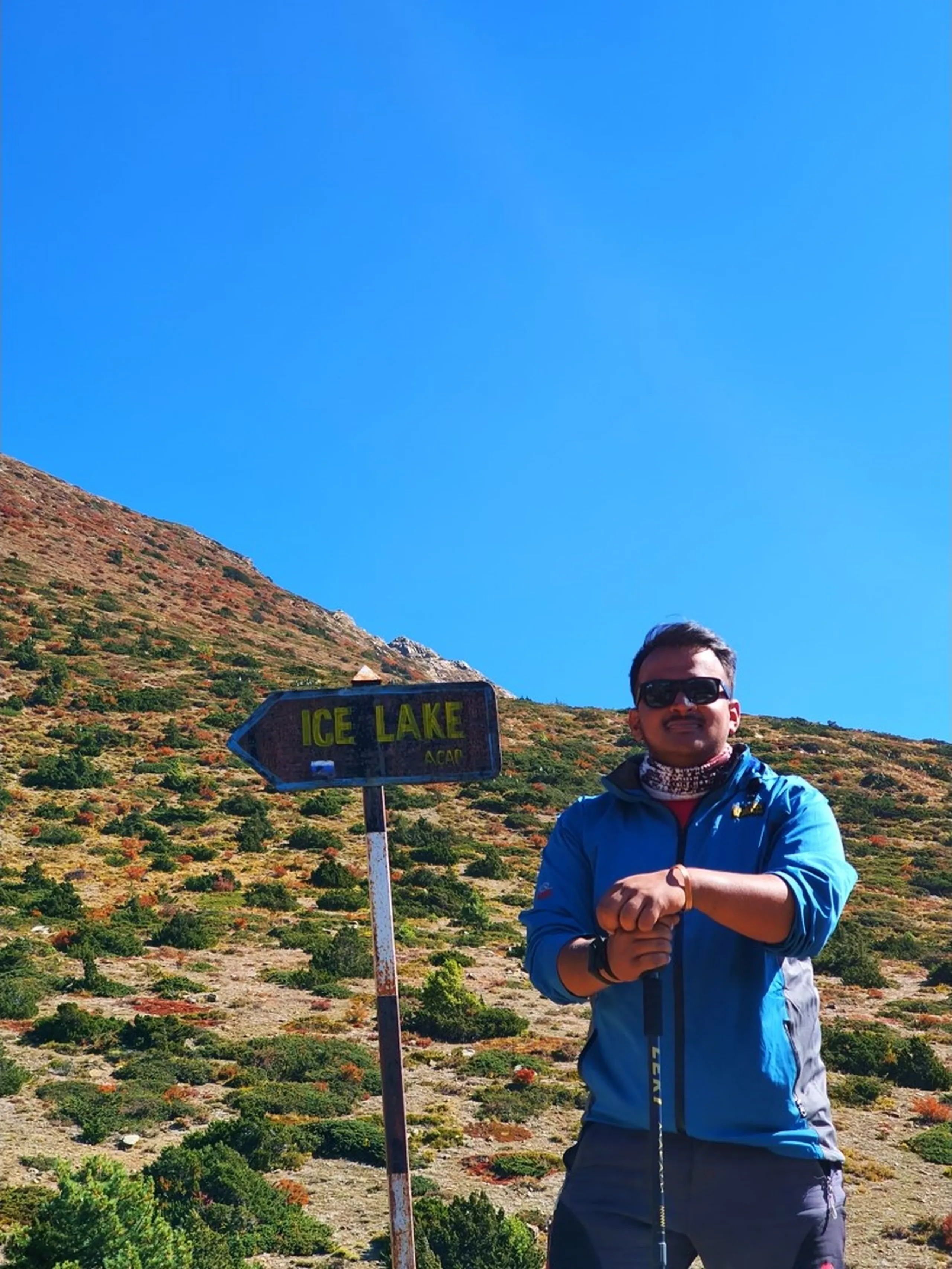 Male trekker in a blue Jack Wolfskin puffer jacket and sunglasses smiling and pointing at a weathered "Ice Lake  ACAP" directional trail sign on a steep scrubby hillside above Manang Nepal, the slope covered in autumn-red and green alpine shrubs leading up to a bare rocky summit, clear blue sky above - Ice Lake Manang Nepal signpost, ACAP trail marker Annapurna Circuit, Ice Lake day hike Nepal