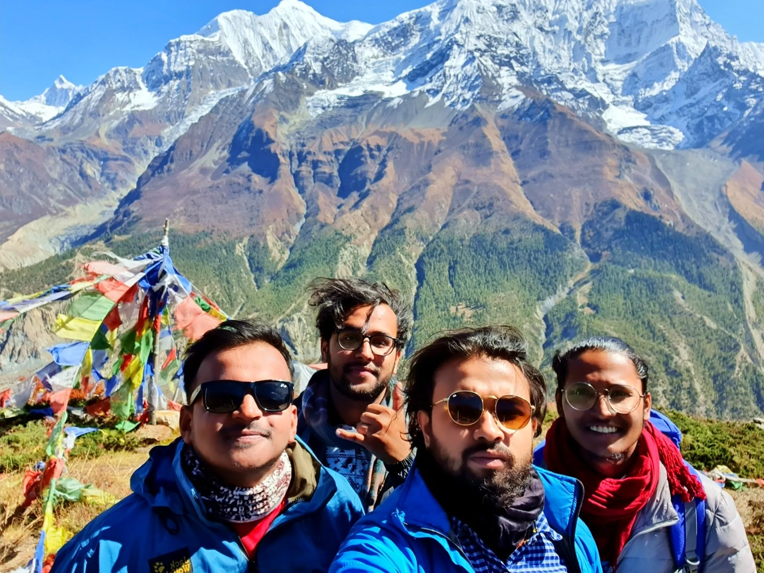 Close-up group selfie of four male trekkers on the Ice Lake trail above Manang Nepal, all wearing sunglasses and colourful trekking jackets in blue, grey and checked patterns, colourful Tibetan Buddhist prayer flags fluttering behind them, and the enormous Annapurna II snowfield with rust-red and pine-forested lower slopes completely filling the background under clear blue sky - Ice Lake Manang group selfie Nepal, Annapurna Circuit trekking friends, high altitude viewpoint Nepal