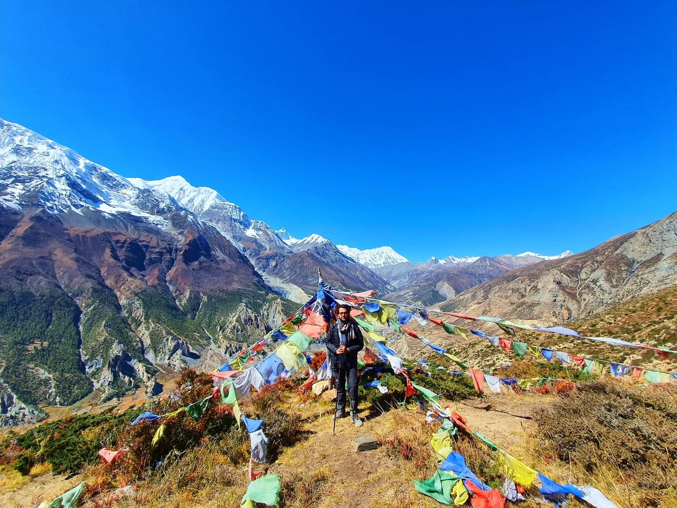 Wide-angle landscape portrait of a lone male trekker in a dark jacket standing amid a dense field of multicoloured Tibetan Buddhist prayer flags on a high ridgeline above Manang Nepal, hundreds of flags in red, green, blue, white and yellow radiating outward from a central pole; the full Annapurna and Gangapurna snowfields spanning the entire left horizon, a deep glaciated valley between them, arid ochre hills to the right under deep blue sky - Ice Lake viewpoint Nepal prayer flags, Annapurna Circuit prayer flag ridge, best viewpoint Manang Nepal