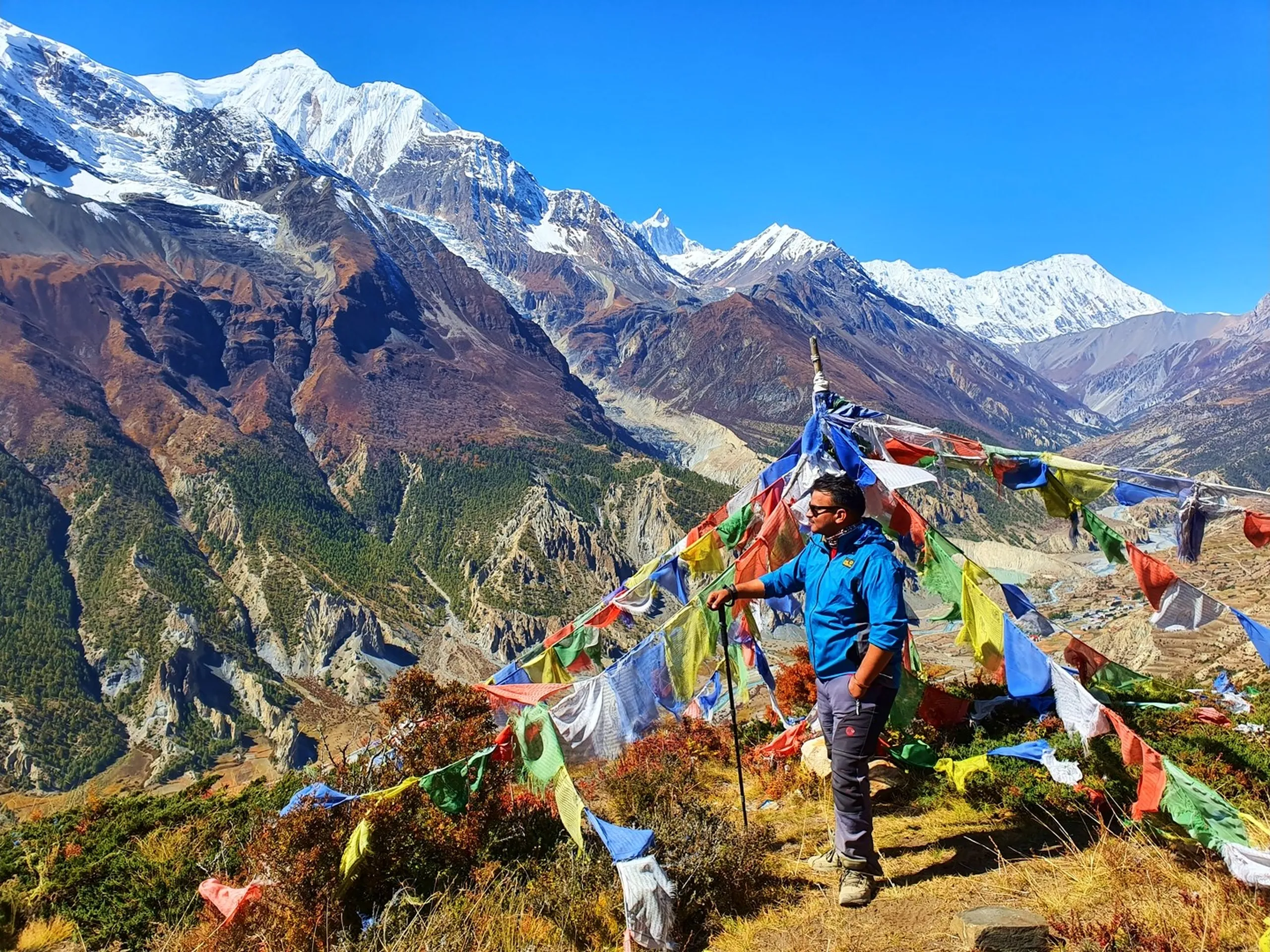 Male trekker in a blue Jack Wolfskin softshell jacket standing confidently with a trekking pole amid a dense array of colourful Tibetan prayer flags on the Ice Lake ridge above Manang Nepal, the full panorama of Annapurna II and Gangapurna snowfields stretching from left to right above rust-red and pine-forested lower ridges, the deep Marsyangdi valley visible below, Manang village visible to the right - Annapurna Circuit prayer flag viewpoint, Ice Lake ridge Nepal, Himalayan panorama trekking Nepal