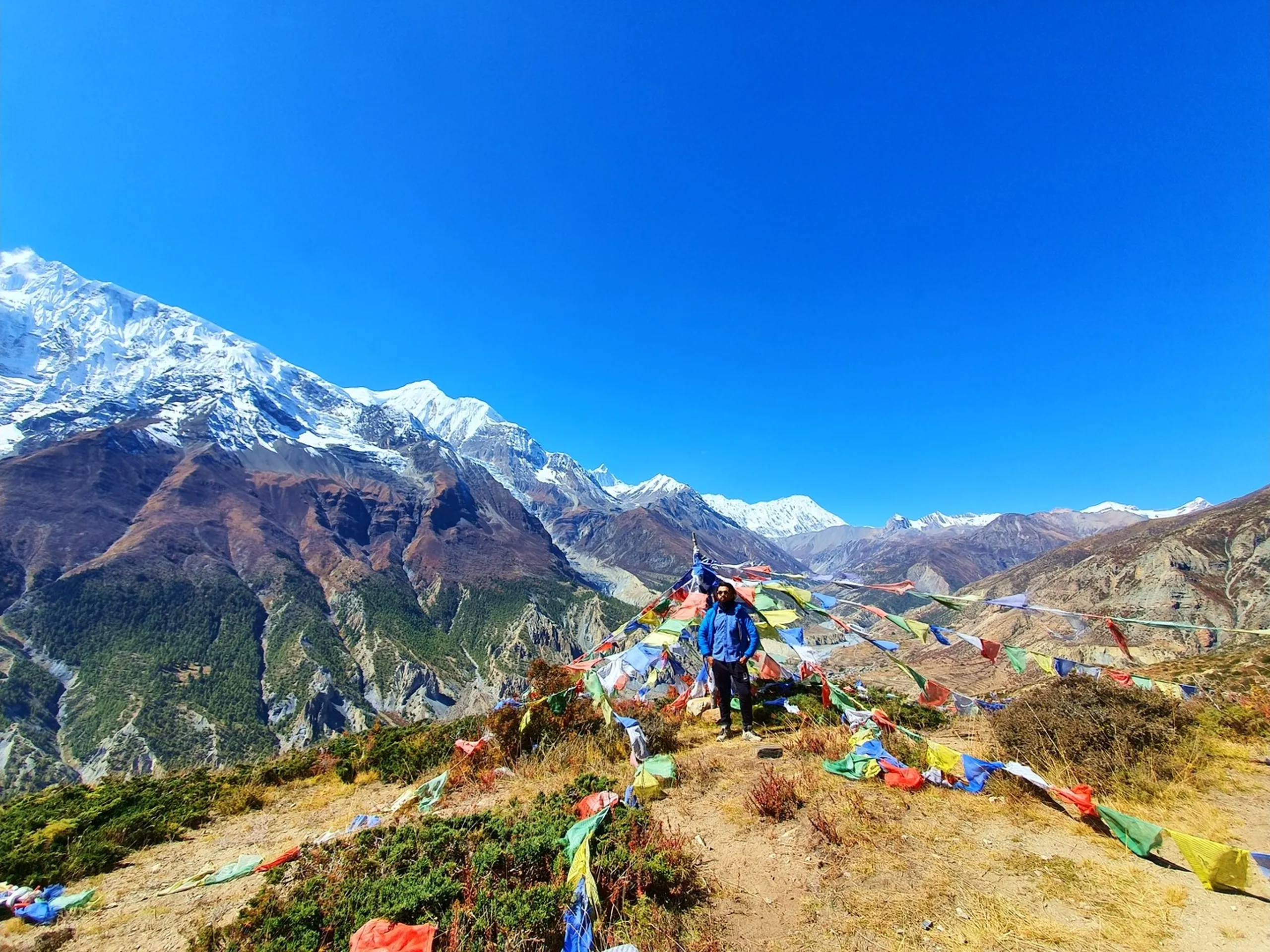 Male trekker in a blue puffer jacket standing on a high bare ridge surrounded by scattered colourful prayer flags in Manang Nepal, the massive Annapurna II snowfield on the far left, a long snow-capped mountain ridgeline extending across the horizon to the right above arid brown valleys, deep blue cloudless sky filling the top half of the frame - Ice Lake day hike ridge Manang, Annapurna panorama Nepal, high altitude trek viewpoint Nepal