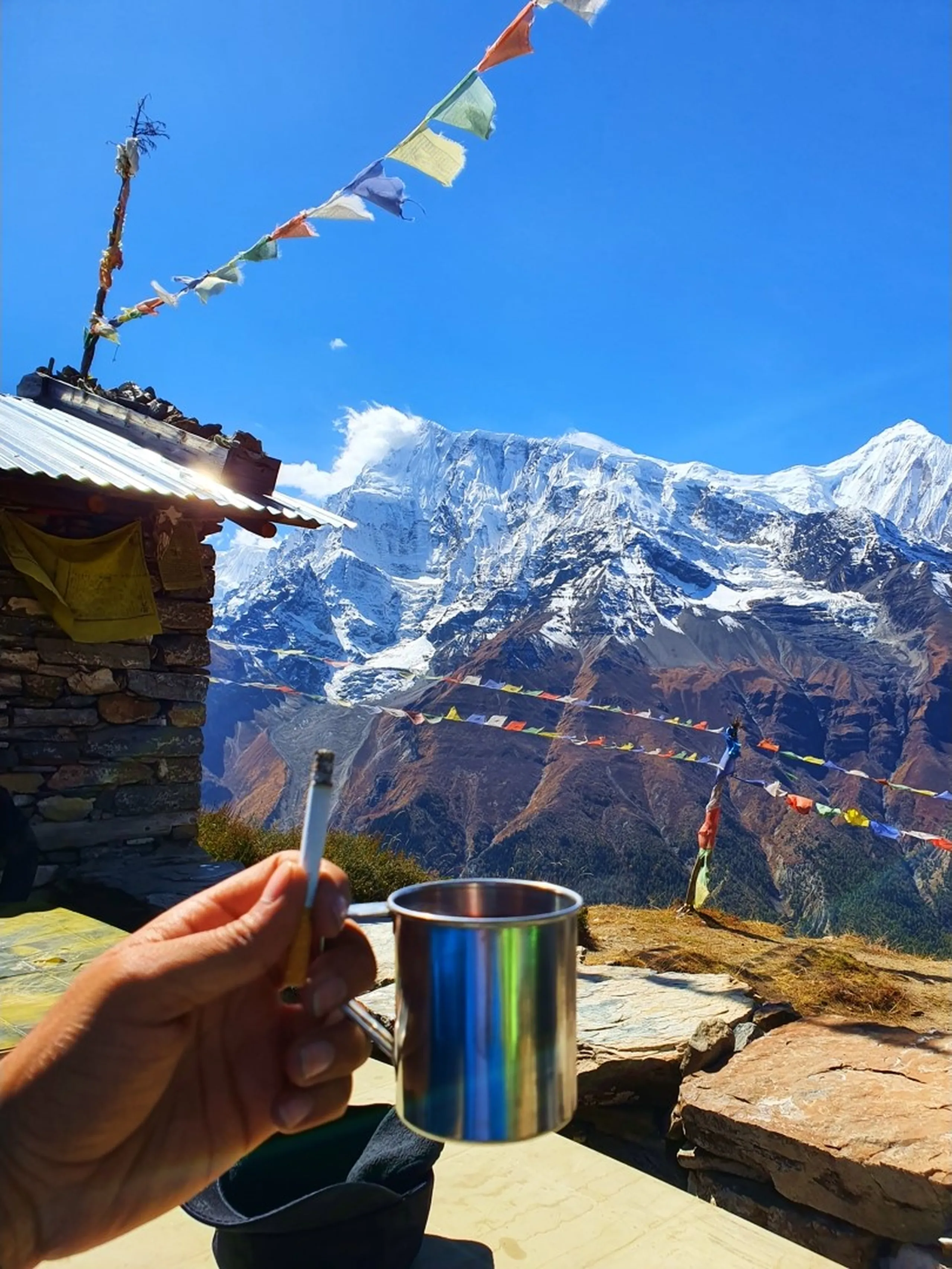 First-person POV of a hand holding a shiny steel camping mug and a cigarette at a tiny stone teahouse on the Ice Lake trail above Manang Nepal, colourful prayer flags strung above, another trekker visible in the distance standing among prayer flags, and the magnificent Annapurna snowfields filling the entire background above forested ridges under brilliant blue sky - tea with Annapurna view Nepal, high altitude tea stop Manang, best view Nepal tea house