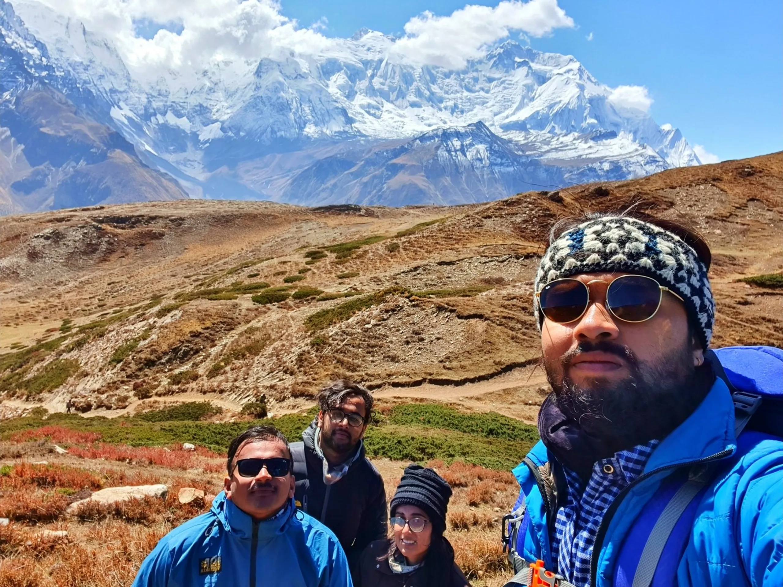 Trekking group selfie with four members on the high-altitude Ice Lake trail in Manang Nepal, one bearded member in a blue Honor puffer jacket and patterned headband taking the selfie in the foreground, three companions visible behind including a woman in a black beanie, another in a black jacket with glasses, and a third in a Jack Wolfskin blue jacket, the Annapurna glacier and snowfield towering dramatically above rolling autumn tundra - Annapurna Circuit group selfie Nepal, Ice Lake trail trekking friends, Manang Nepal family trek