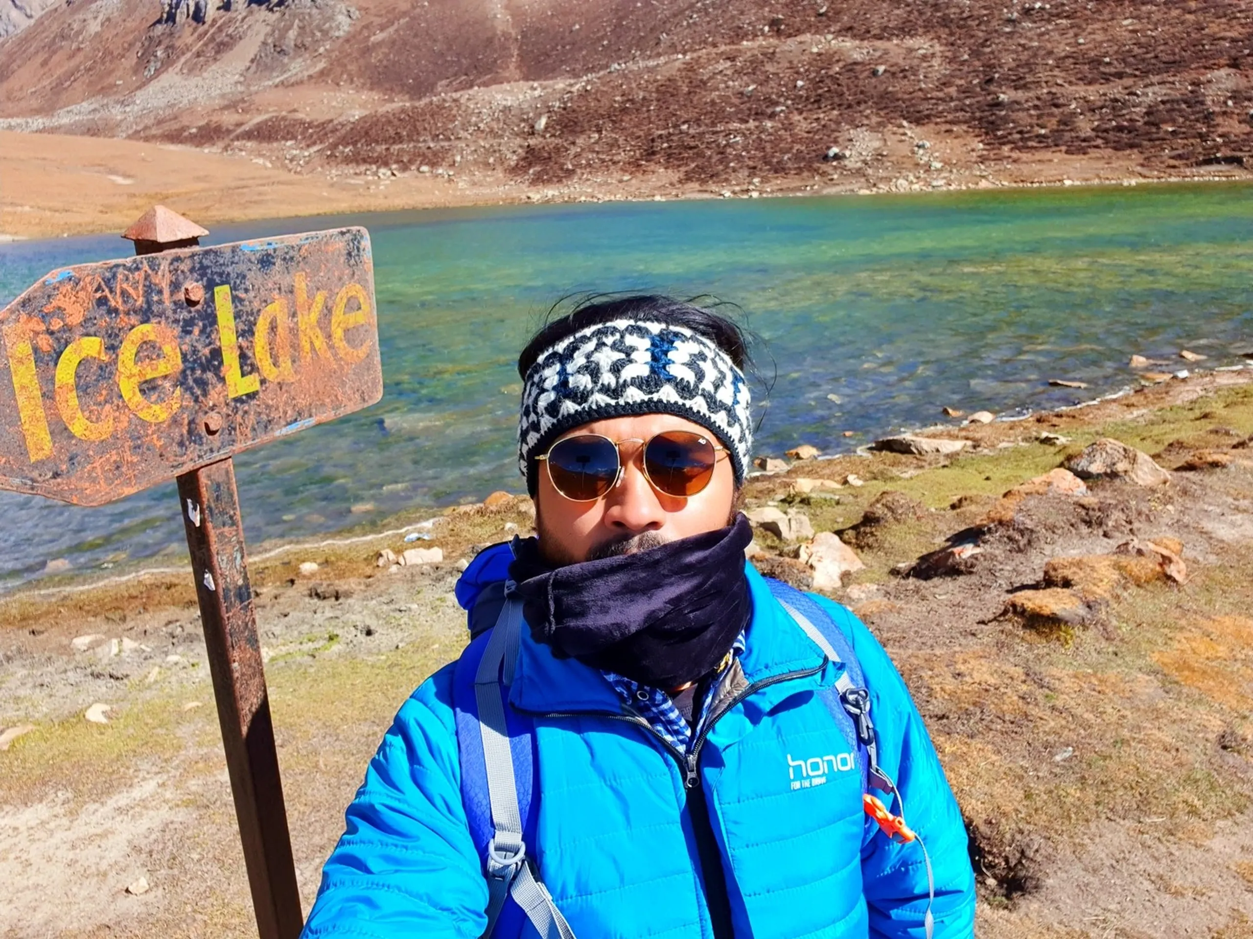 Male trekker in a blue Honor puffer jacket and patterned headband with amber round sunglasses and dark neck gaiter, standing beside a rusty weathered "Ice Lake" metal sign on a post at the actual shore of Ice Lake Nepal, the brilliant emerald-green and turquoise glacial lake surface visible directly behind him, bare brown rocky slopes rising beyond, achievement expression on face - Ice Lake Nepal reached, Annapurna Circuit Ice Lake sign, 4600m high altitude lake Nepal