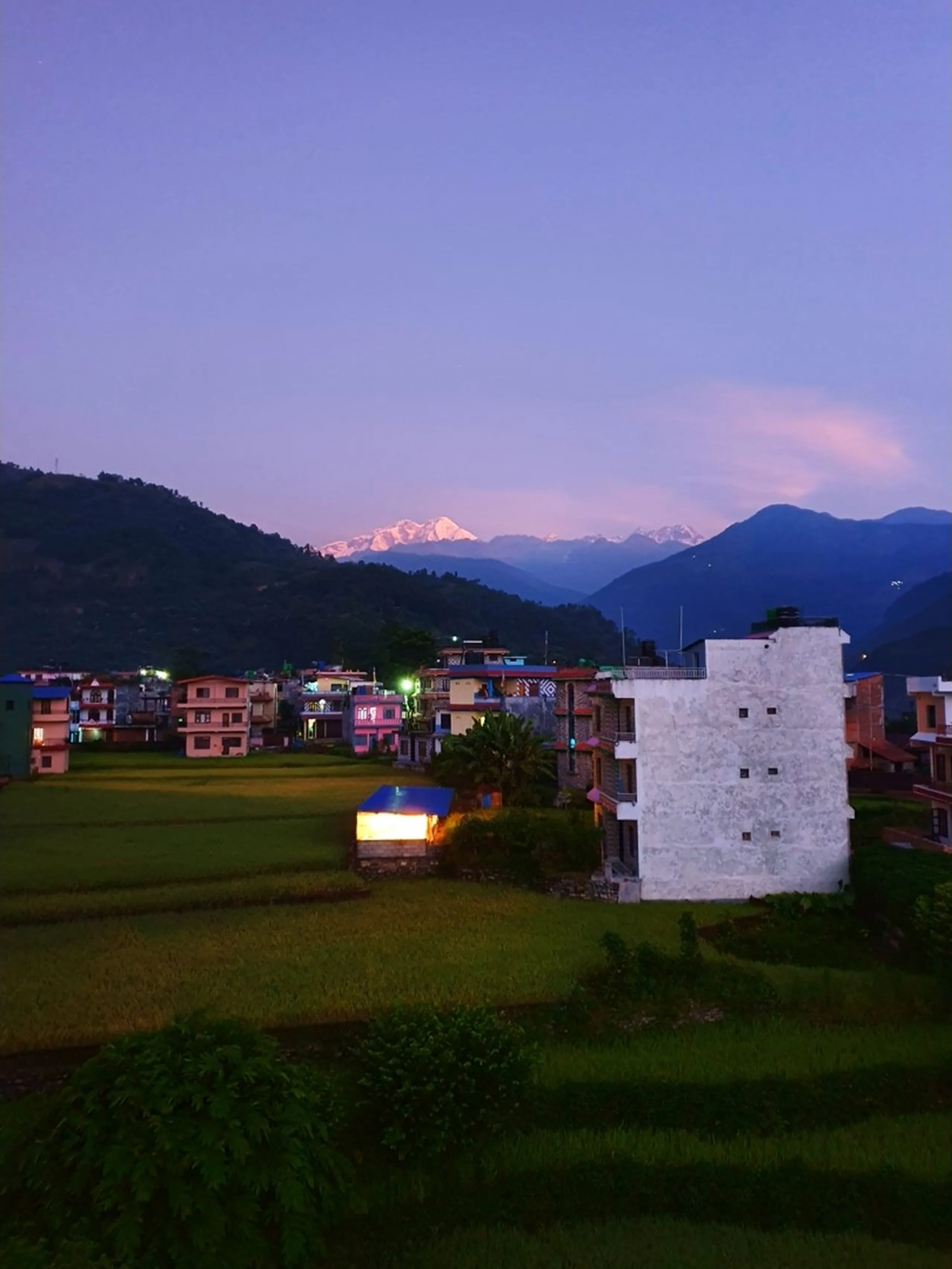 A breathtaking twilight panorama from a resort balcony in the Lamjung district of Nepal - lush green paddy rice fields glow in the last light of dusk, a traditional Nepali village with colorful homes and warm glowing windows nestled in the middle ground, and in the far distance the snow-capped peaks of the Annapurna Himalayan range glow in alpenglow pink and white against a deep violet-blue sky with a single pink cloud drifting above - a stunning Nepal mountain sunrise and rice terrace landscape ideal for Annapurna Circuit travel blogs and Nepal Himalaya photography