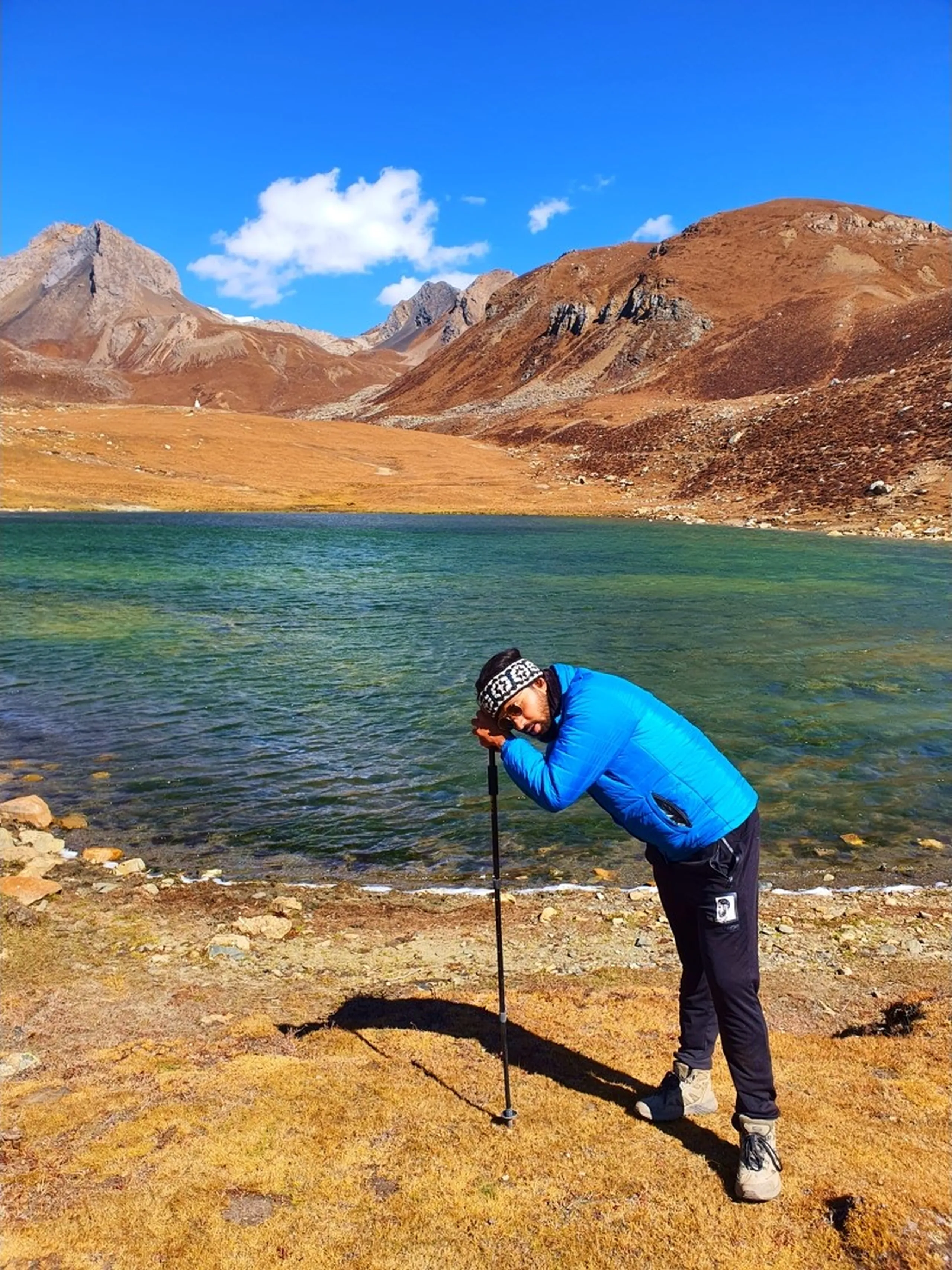 Male trekker in a blue puffer jacket and patterned headband bent over and leaning exhausted on a trekking pole at the rocky shore of Ice Lake Nepal at 4600 metres, the vivid emerald-green transparent glacial lake stretching behind him, bare rust-brown rocky mountains reflected in the still water, a single white cloud in the deep blue sky - exhausted trekker Ice Lake Nepal, altitude fatigue 4600m Annapurna Circuit, Ice Lake Manang achievement