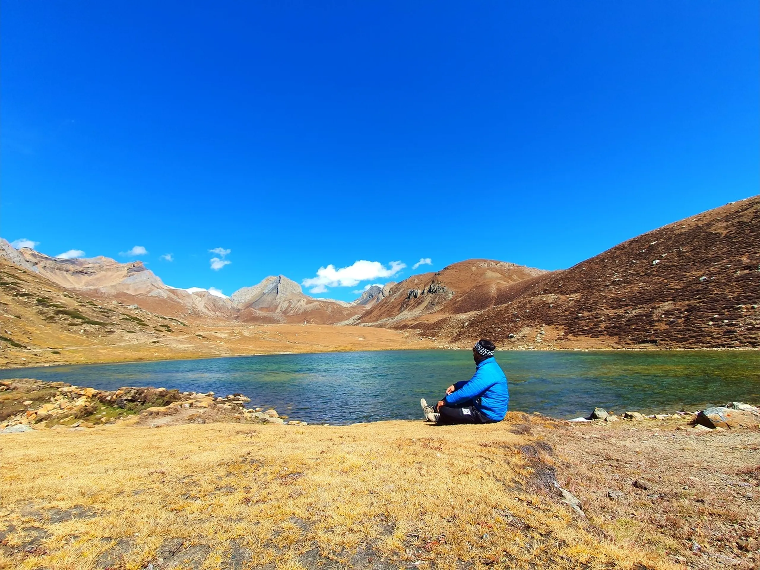 Solitary trekker in a blue puffer jacket sitting cross-legged on golden dried grass at the shore of Ice Lake Nepal at 4600 metres, gazing contemplatively at the vivid emerald-green glacial lake, wide open landscape of bare ochre mountains encircling the lake on all sides, deep blue sky with a single cloud - Ice Lake Manang Nepal meditation, solo trekker high altitude lake Nepal, peaceful Himalayan lake shore