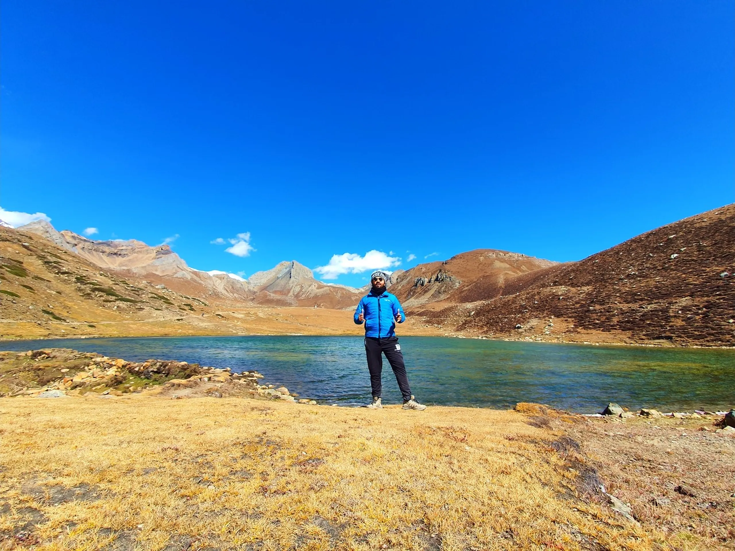 Male trekker in a blue puffer jacket and patterned headband giving two thumbs-up and standing at the shore of Ice Lake Nepal at 4600 metres altitude, the vivid emerald-green transparent glacial lake behind him, bare rust-brown rocky peaks encircling the alpine bowl, small white cloud in the electric blue sky - Ice Lake Nepal achievement thumbs up, Annapurna Circuit Ice Lake 4600m, Nepal high altitude trek success
