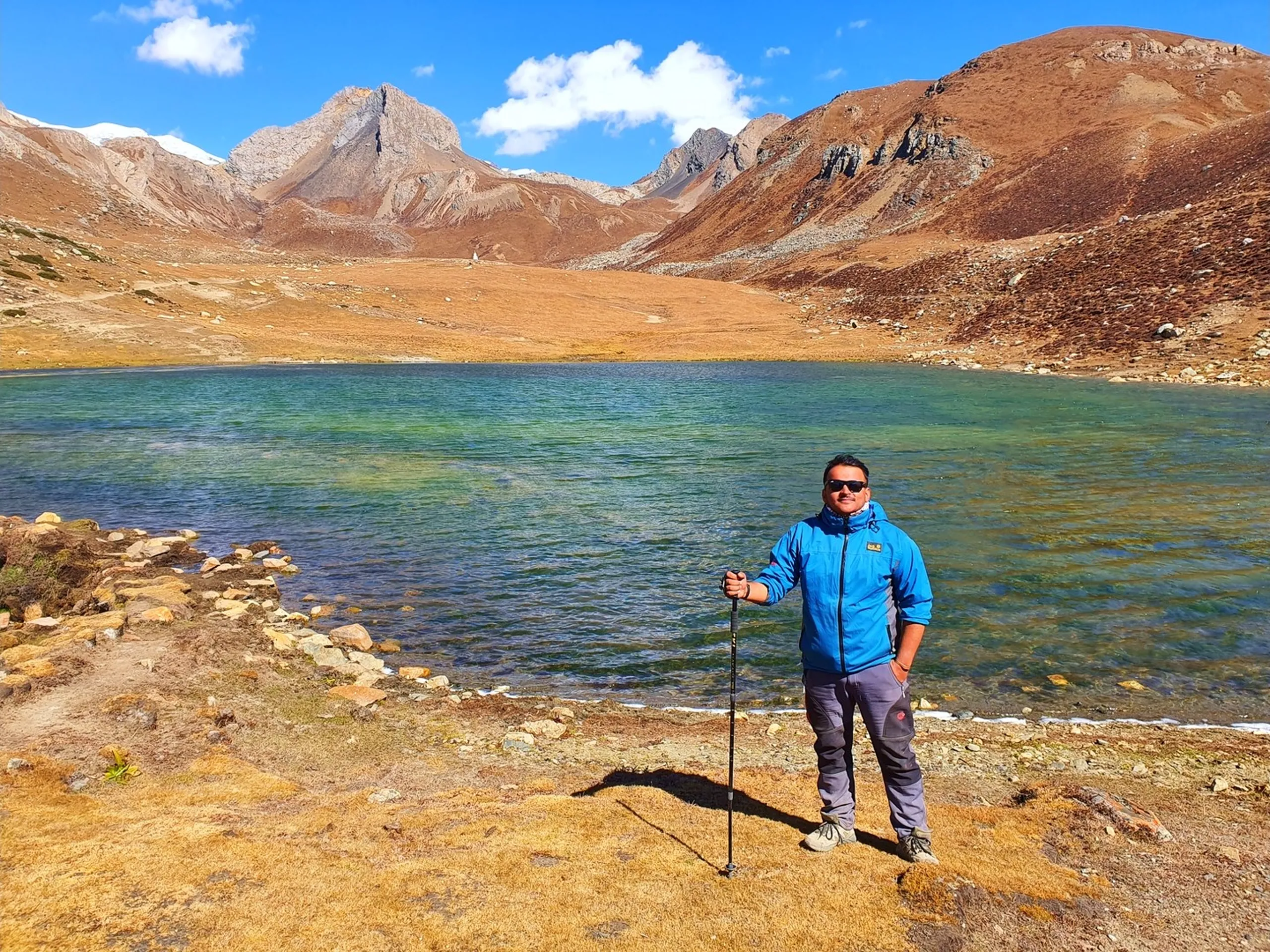 Male trekker in a blue Jack Wolfskin softshell jacket holding a trekking pole at the rocky pebbly shore of Ice Lake Nepal at 4600 metres, the vivid emerald-green and turquoise glacial lake spreading behind him, rugged grey and rust mountain ridges rising on both sides with a small stupa visible on a distant ridge, under blue sky with white clouds - Ice Lake Manang Nepal 4600m, Annapurna Circuit high altitude lake, trekker at Ice Lake Nepal