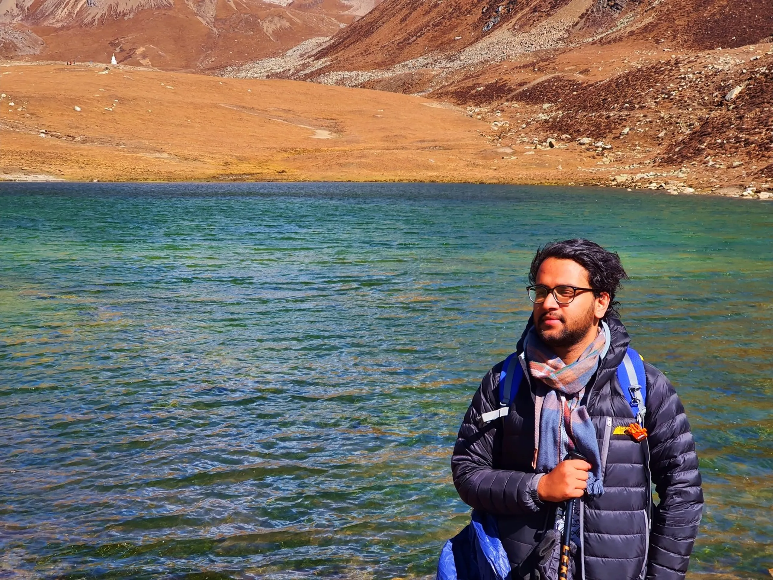 Contemplative male trekker in a dark charcoal puffer jacket and plaid scarf with black-framed glasses standing at the shore of Ice Lake Nepal at 4600 metres, looking to the side with a wistful expression, the vivid blue-green glacial lake rippling in the foreground, golden alpine tundra and a small white stupa on a distant ridge visible behind, rust-brown mountain slopes under partially cloudy blue sky - Ice Lake Nepal contemplative portrait, Annapurna Circuit high altitude lake, Manang Ice Lake trekker