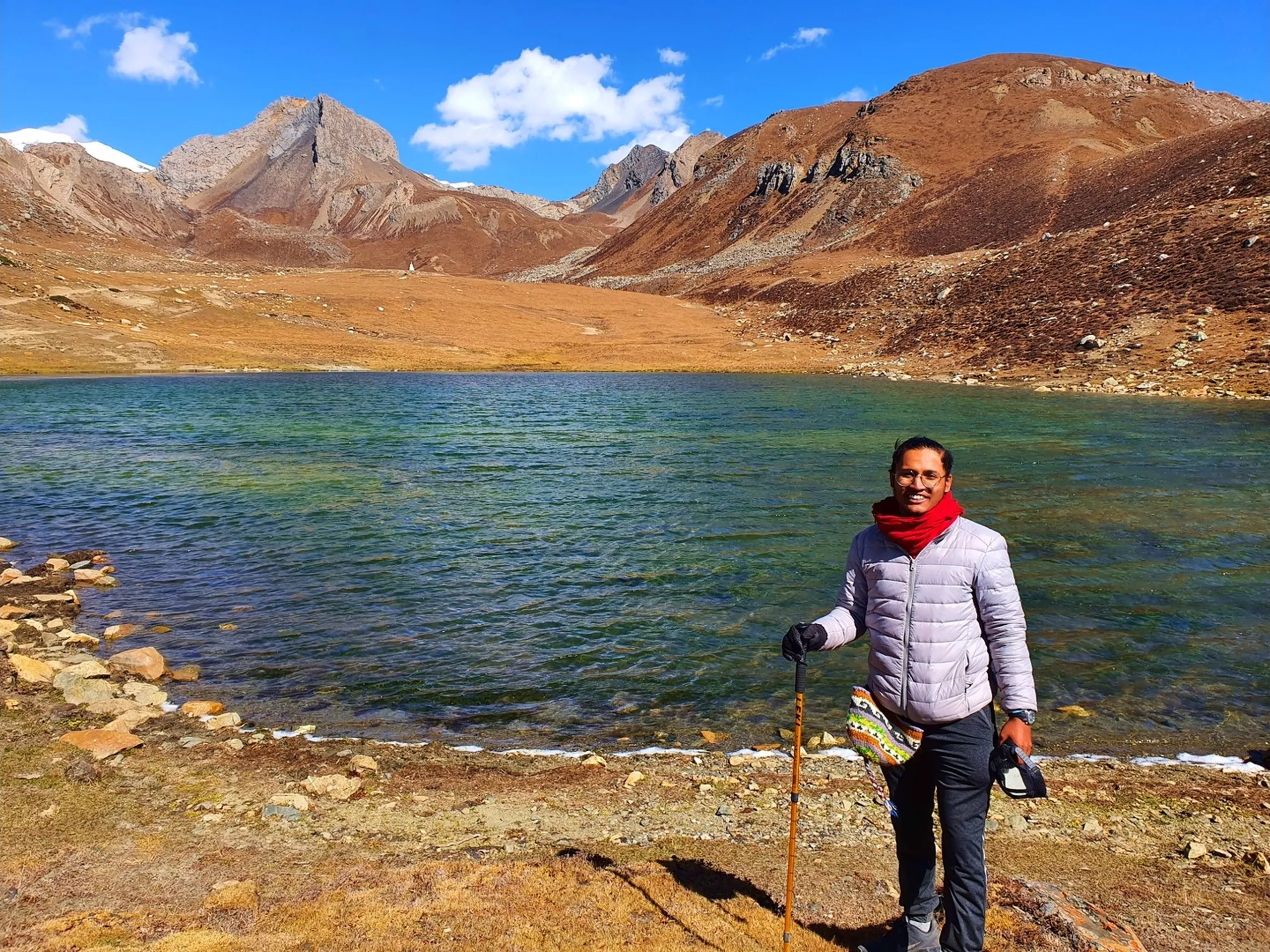 Smiling male trekker in a silver-grey puffer jacket and red scarf holding a trekking pole at the shore of Ice Lake Nepal at 4600 metres, the vivid emerald-green glacial lake and bare rust-brown rocky mountains forming a perfect circle behind him, scattered snow patches on the upper slopes, partly cloudy blue sky - Ice Lake Nepal 4600m smiling trekker, Annapurna Circuit Ice Lake, high altitude trekking achievement Nepal