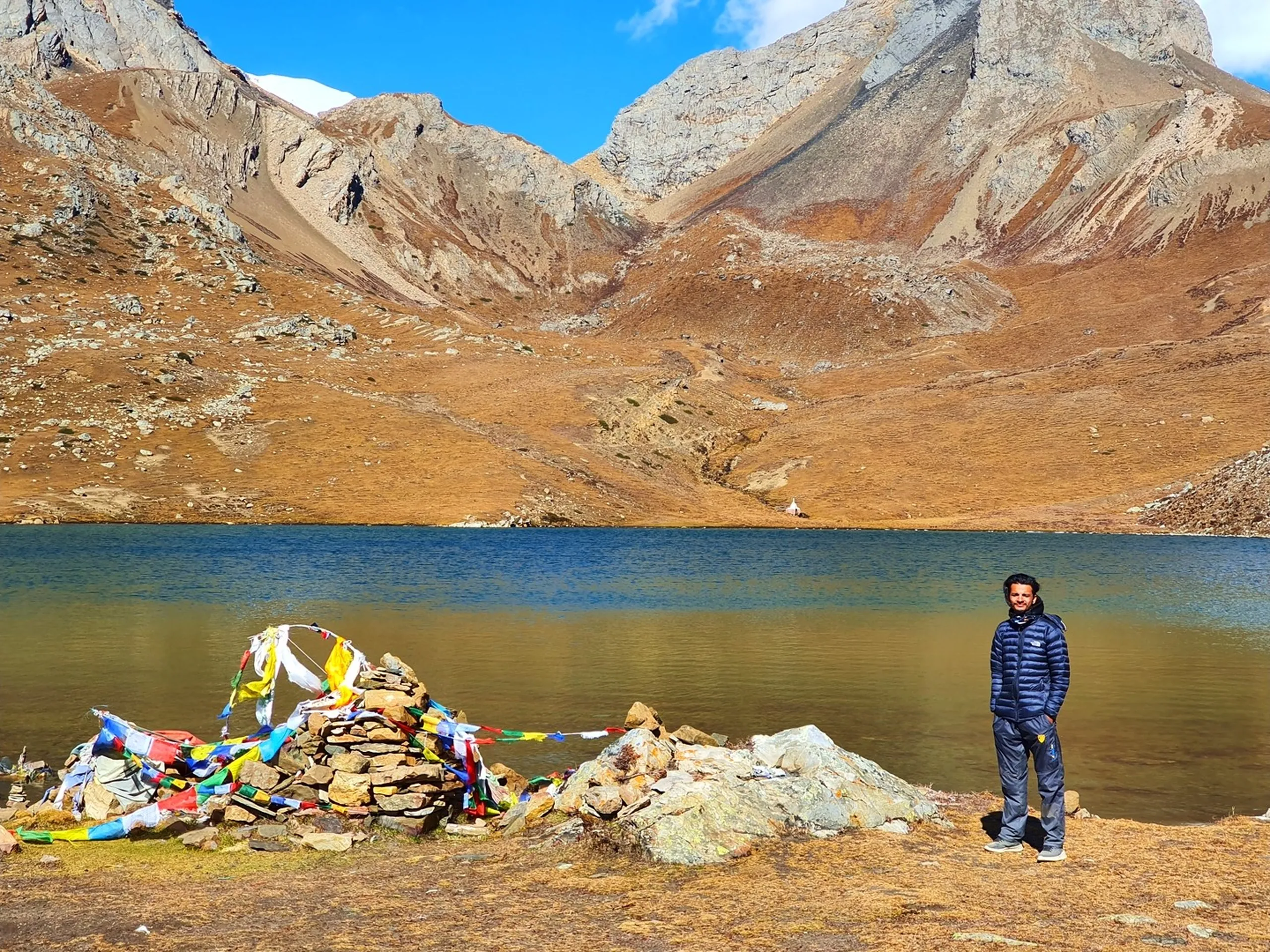 Male trekker in a dark navy blue puffer jacket standing at the shore of Ice Lake Nepal, a stone cairn decorated with multicolour Tibetan prayer flags directly beside him, the vivid blue-green glacial lake reflecting the sky behind, two dramatic grey rocky peaks flanking a high mountain pass in the background, golden alpine meadow and rust-brown slopes - Ice Lake Nepal prayer flag cairn, Annapurna Circuit summit lake, Manang Ice Lake achievement Nepal