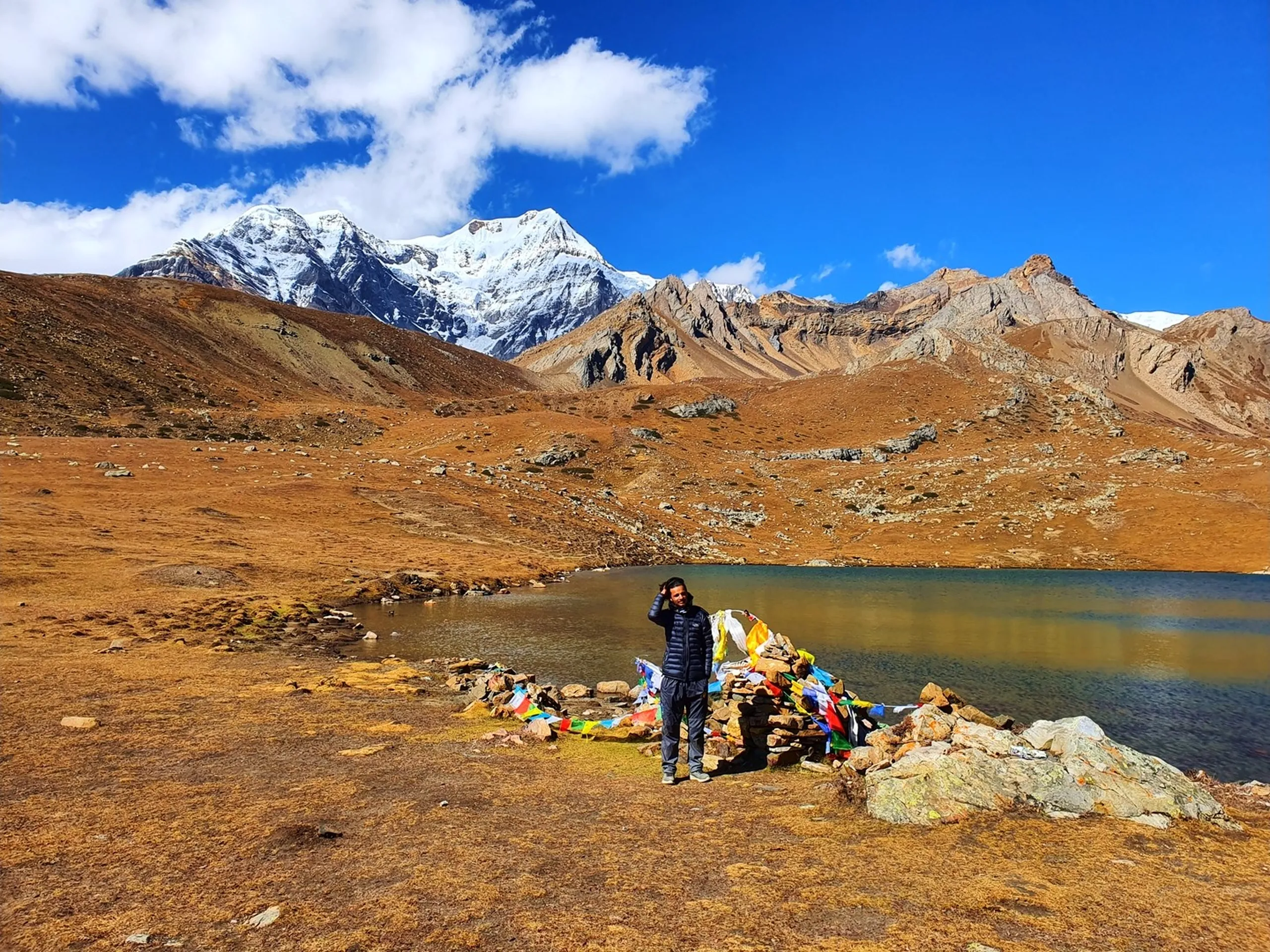 Wide landscape portrait of a male trekker in a dark navy puffer jacket standing beside a colourful prayer flag cairn at Ice Lake Nepal at 4600 metres, the full breath of the scene visible - turquoise glacial lake, rust-brown high alpine tundra, a snow-capped Himalayan peak on the upper left, jagged rocky ridgelines to the right - all under vivid blue sky with white clouds - Ice Lake Manang Nepal landscape, Annapurna Circuit 4600m lake, best lake trek Nepal