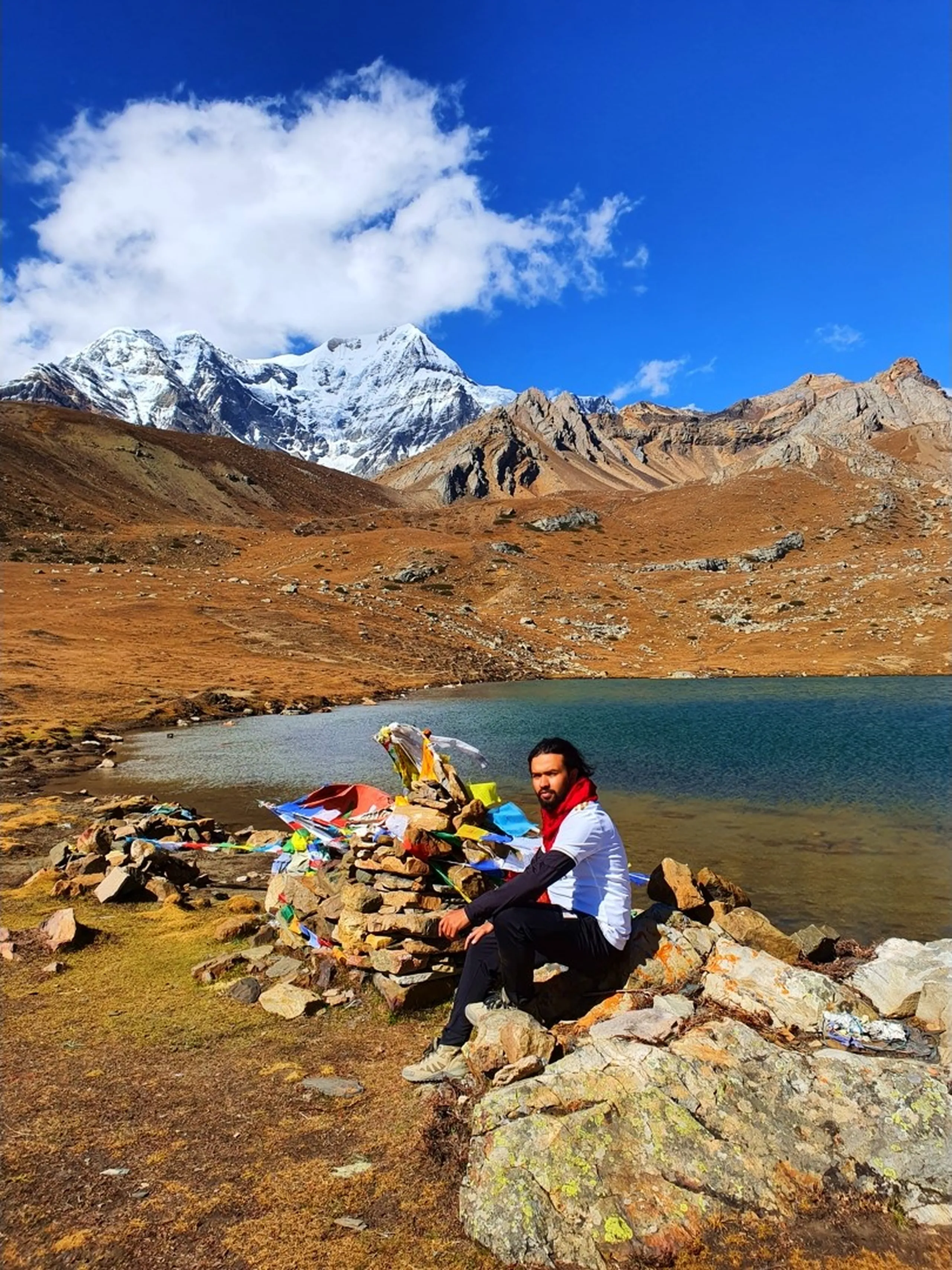 Male trekker with long dark hair in a white long-sleeve shirt and red scarf sitting relaxed on a stone cairn decorated with colourful Tibetan Buddhist prayer flags at Ice Lake Nepal at 4600 metres, the blue-green glacial lake visible behind him, a snow-capped peak on the upper left and jagged rocky ridges to the right, golden autumn alpine tundra all around - Ice Lake Nepal relaxed trekker, Annapurna Circuit cairn prayer flags, 4600m high altitude Nepal