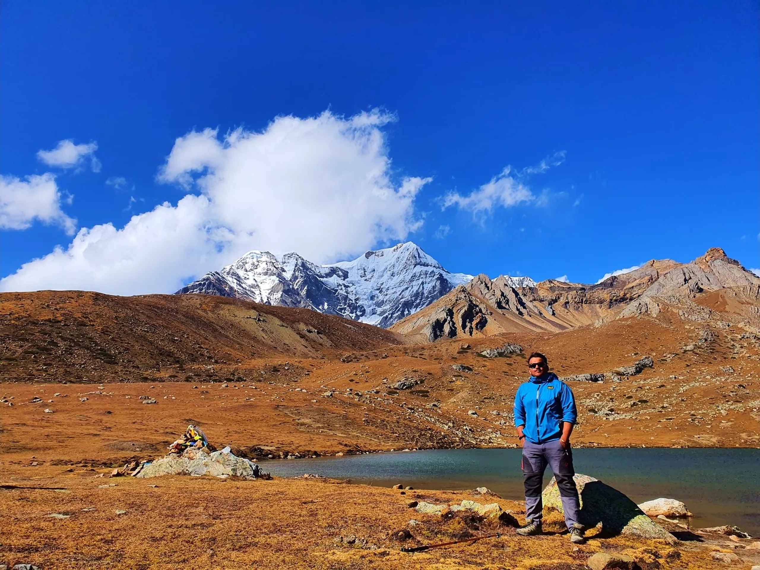 Male trekker in a blue Jack Wolfskin softshell jacket and dark sunglasses standing on the golden alpine tundra beside Ice Lake Nepal at 4600 metres, the vivid blue-green glacial lake in the middle ground, a prayer flag cairn to his left, and a dramatic snow-capped Himalayan peak rising directly behind him above arid rocky ridgelines under a vivid blue sky with white clouds - Ice Lake Nepal blue jacket trekker, Annapurna Circuit high lake Nepal, Manang Ice Lake day hike