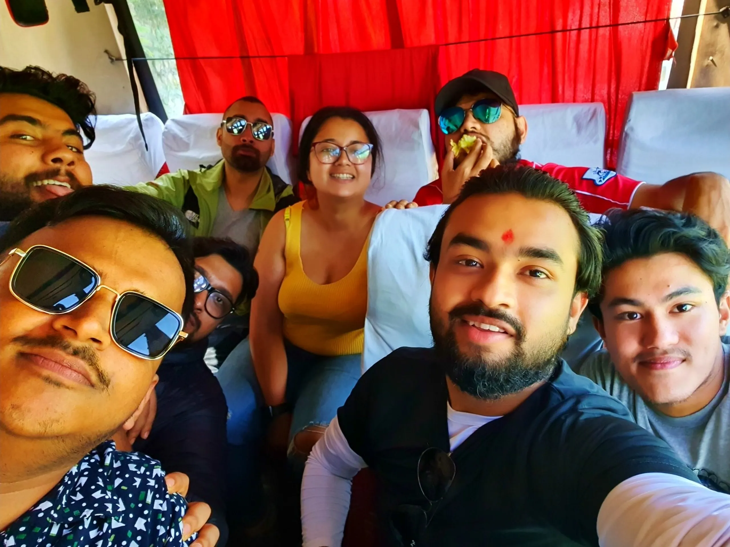 A cheerful group of eight young Nepali trekkers taking a bus selfie on the road to Manang, Nepal - some wearing sunglasses, one with a traditional tika on his forehead, another casually snacking, all laughing and relaxed against the backdrop of red curtain bus seats - a classic Nepal trekking group photo full of friendship and pre-trek excitement